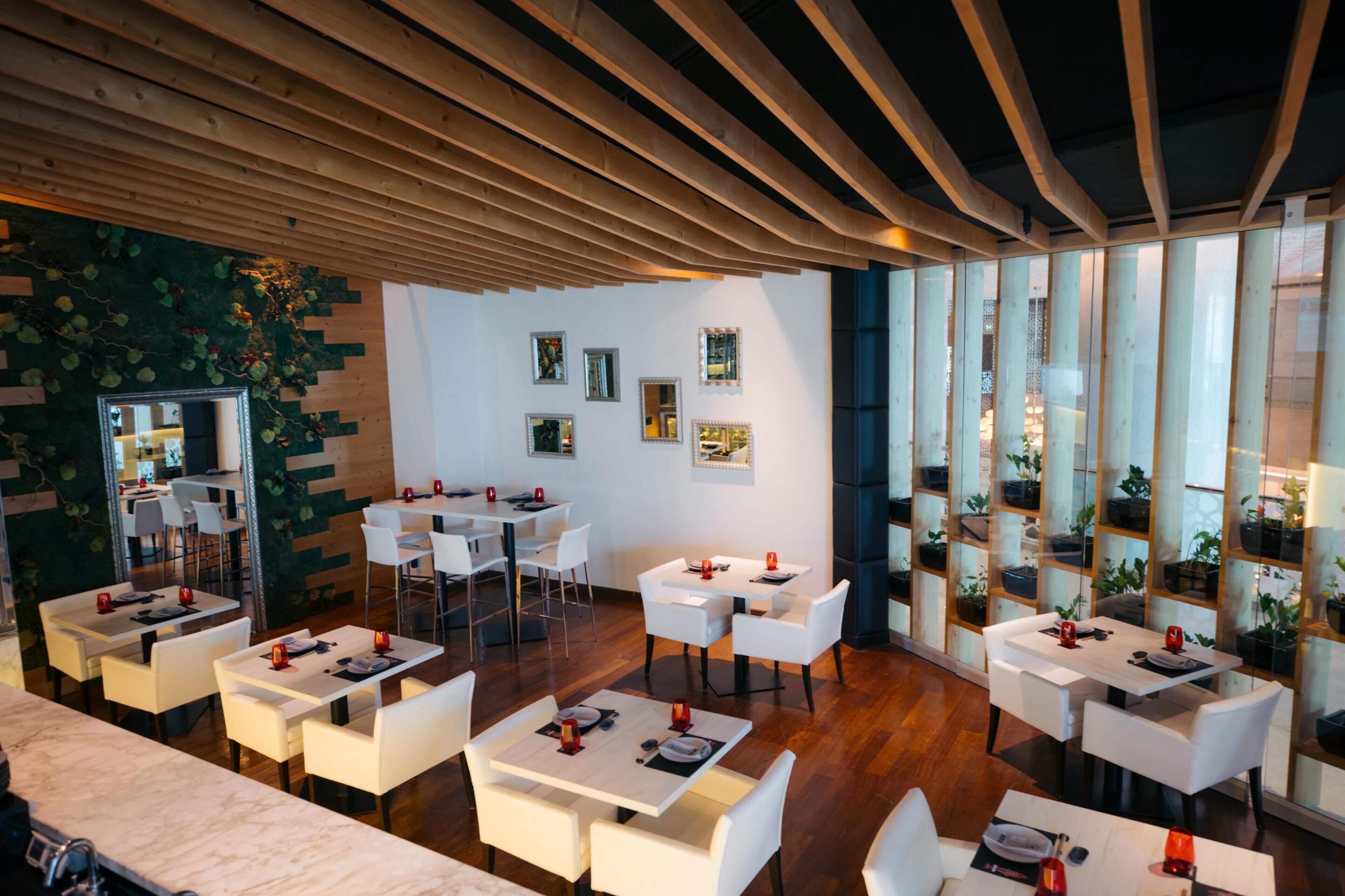 Un restaurant moderne avec des tables et des chaises blanches, un plafond en bois et un mur vert orné de plantes.