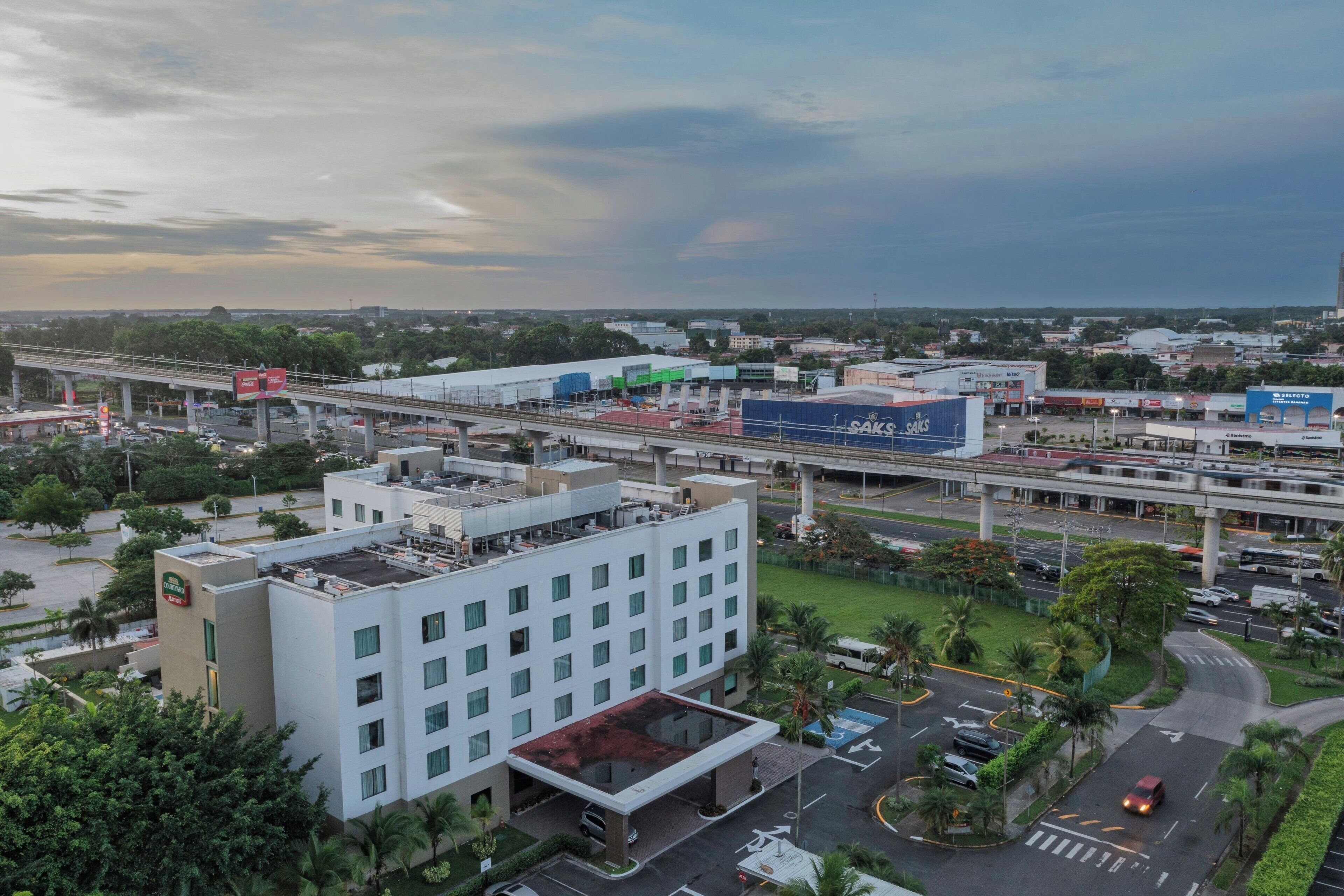 A multi-story hotel with a flat roof, surrounded by greenery and a busy road with an overpass.