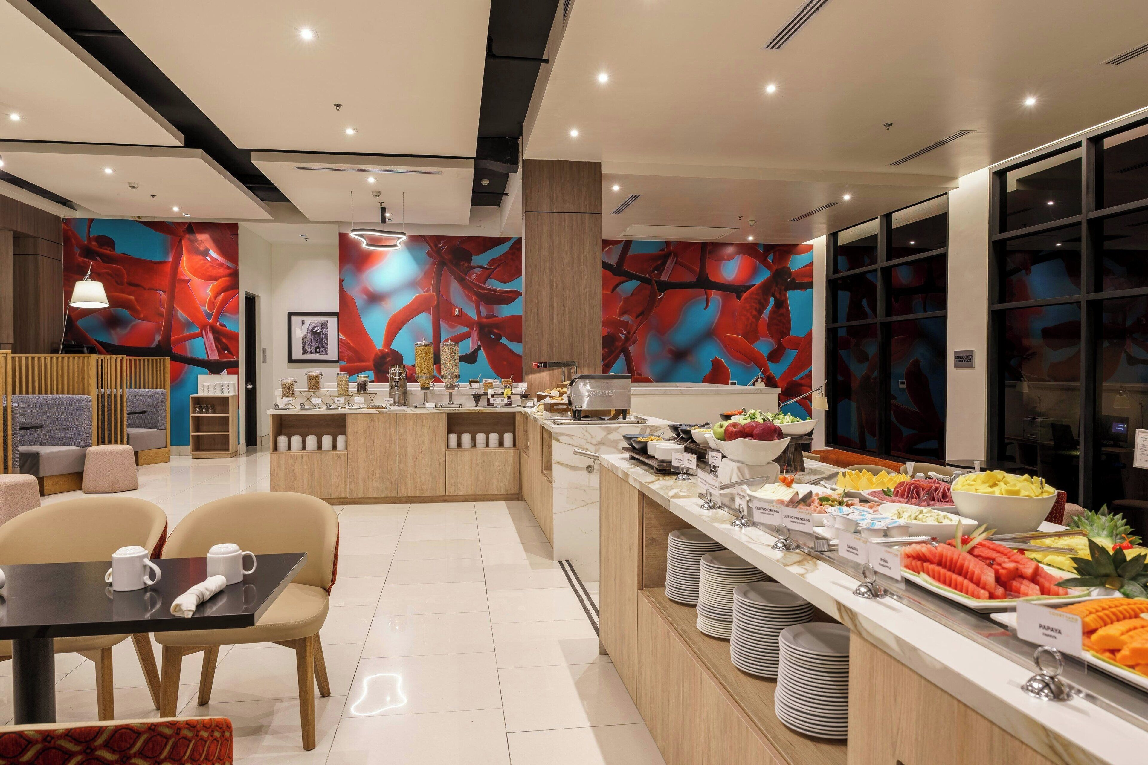 Un moderno hall de hotel con un mostrador central que ofrece una variedad de alimentos para el desayuno, zonas de estar y un gran mural abstracto en las paredes.