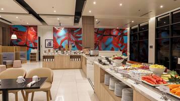 Un moderno hall de hotel con un mostrador central que ofrece una variedad de alimentos para el desayuno, zonas de estar y un gran mural abstracto en las paredes.