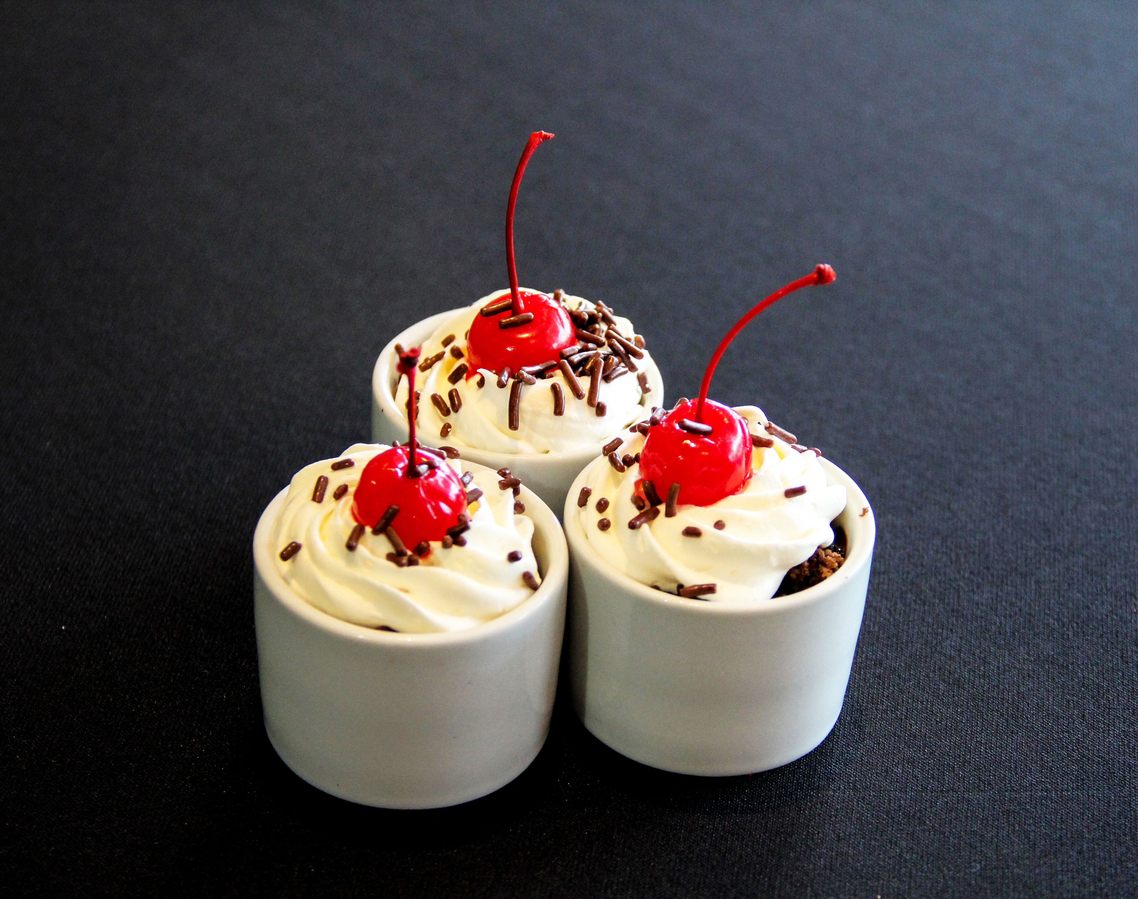 Tres cupcakes con crema batida y cerezas sobre una superficie oscura.