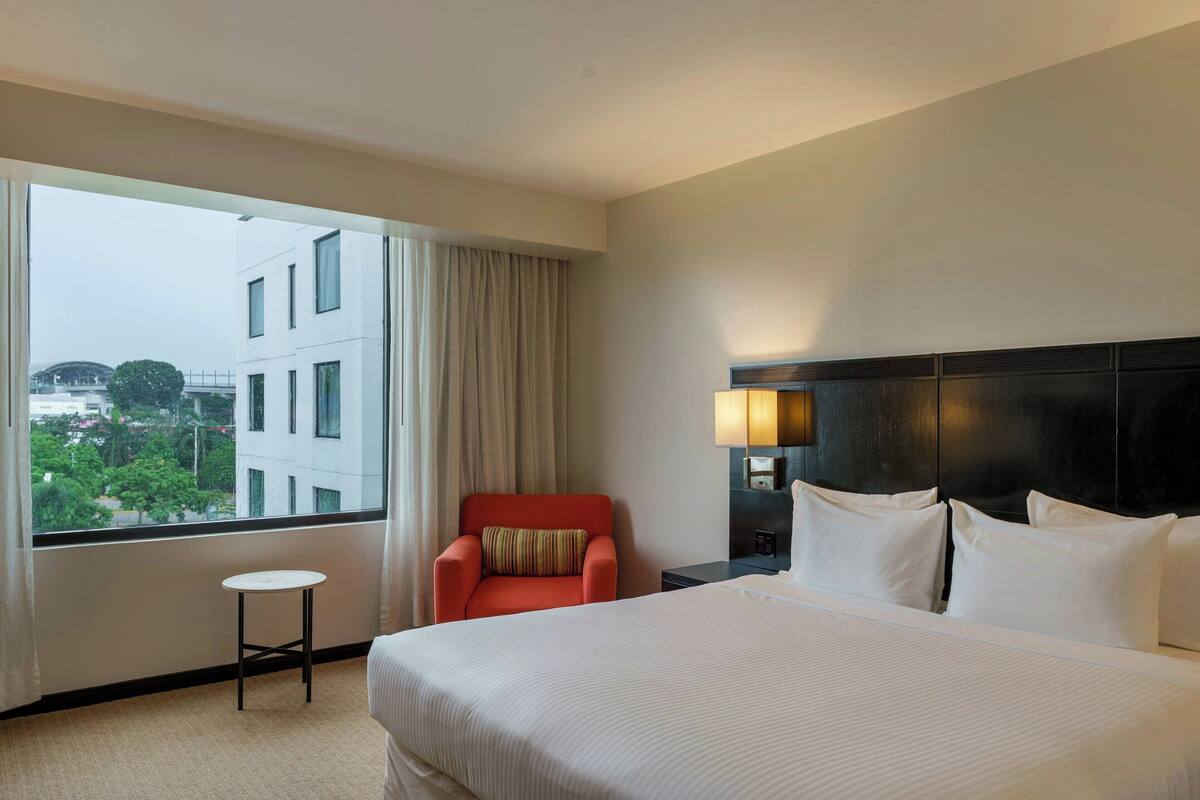 Une chambre d’hôtel avec un grand lit, une petite table, un fauteuil rouge, une table de chevet avec une lampe, et une vue sur les bâtiments et les arbres par la fenêtre.