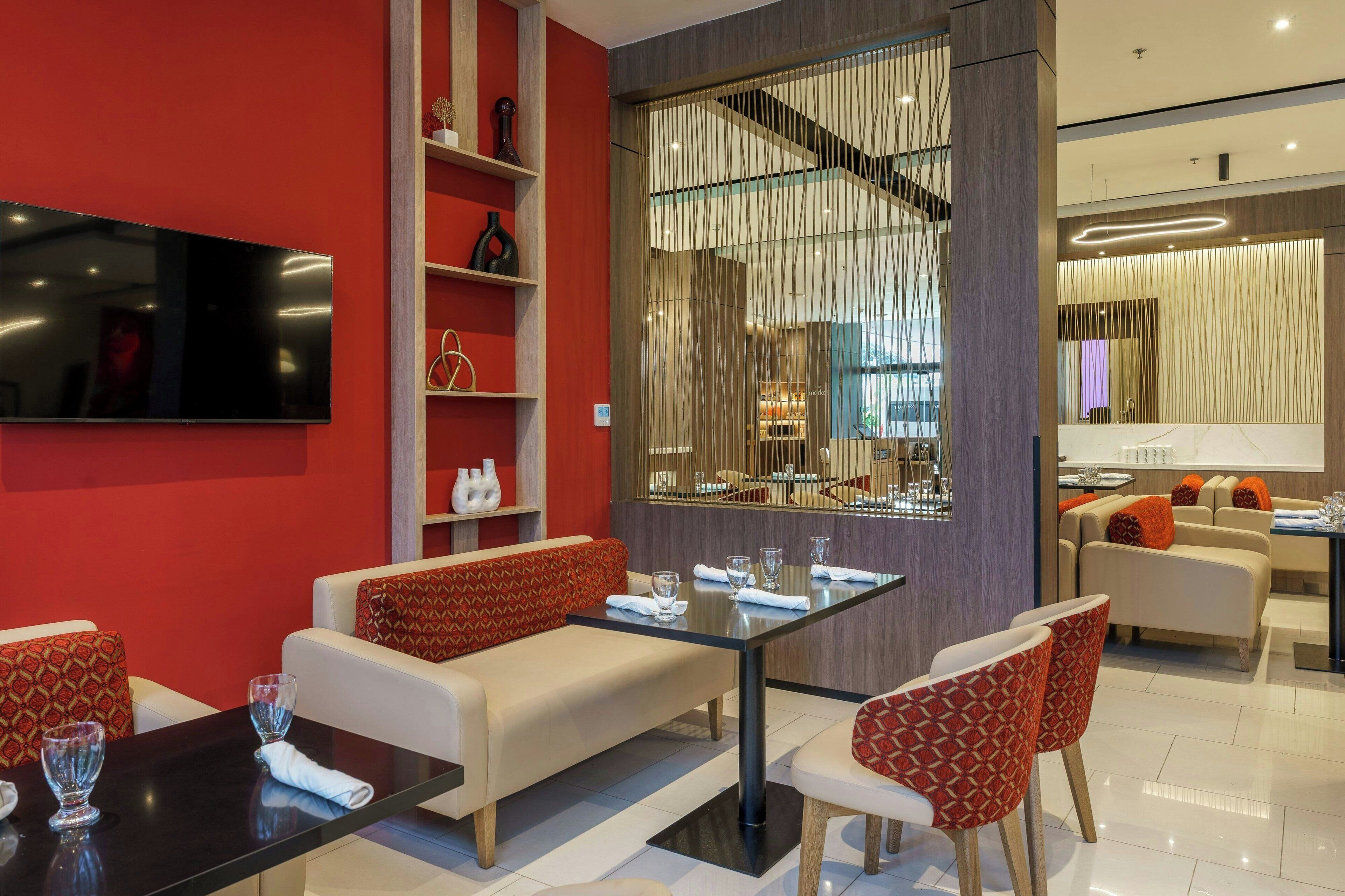 Quarto de hotel moderno com sofá, mesa de jantar e cadeiras. Há uma televisão na parede e uma janela com cortinas.