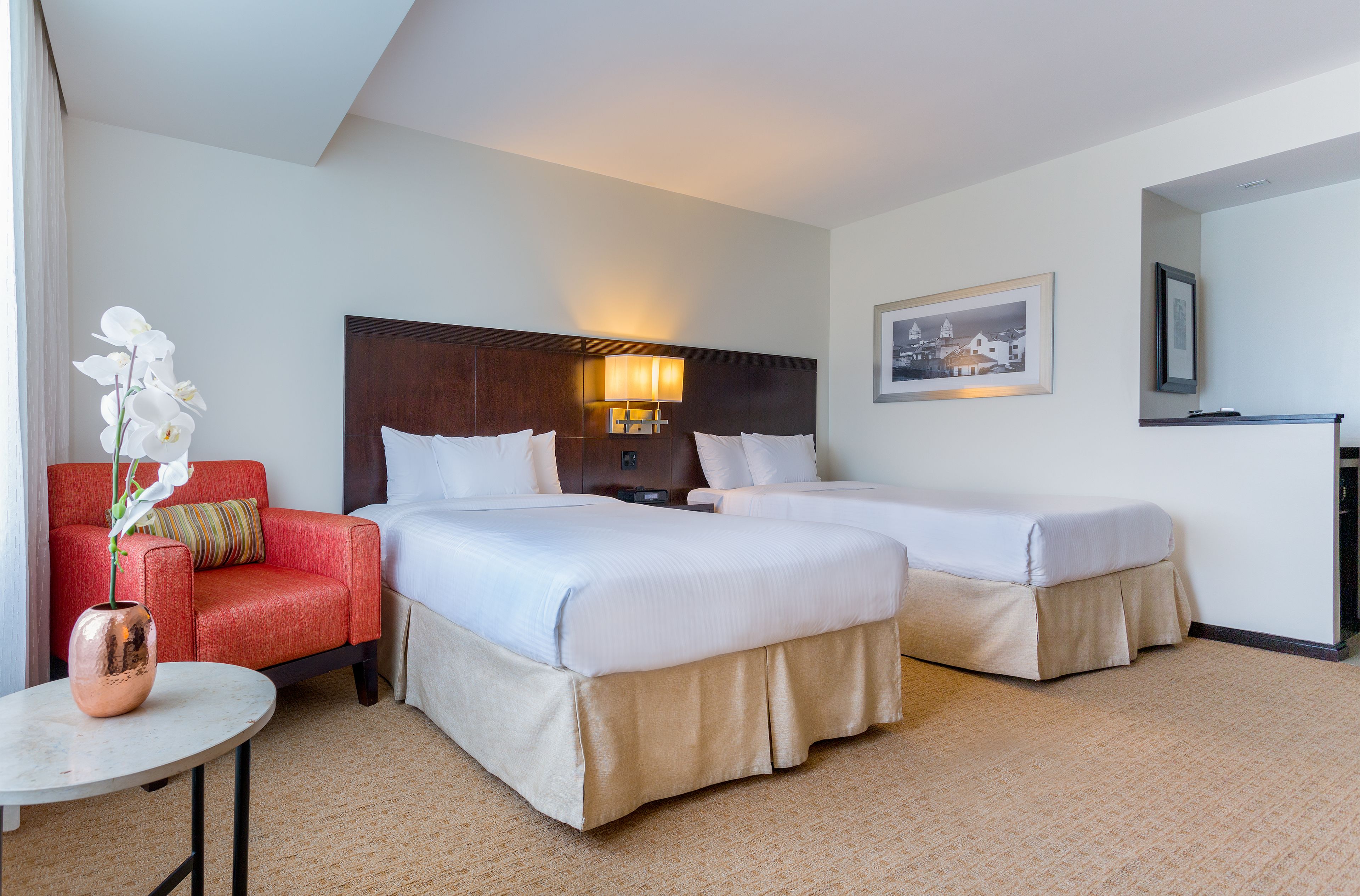 Habitación de hotel con dos camas, un sillón rojo, una mesita con un jarrón y un cuadro en la pared.