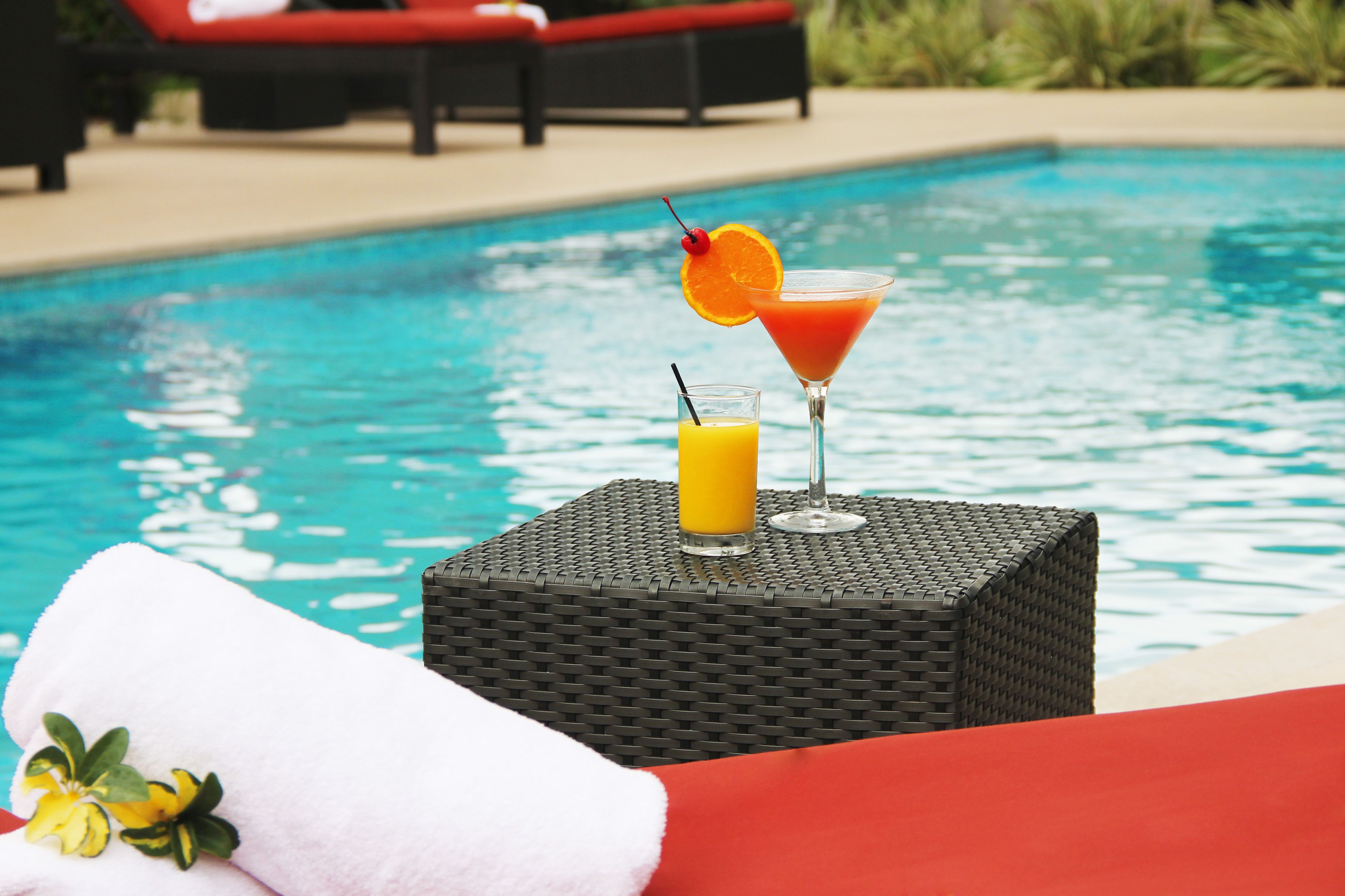 Escena junto a la piscina con una mesa de mimbre que sostiene una copa de martini y un vaso de jugo de naranja, una toalla con estampado floral y una silla de descanso roja al fondo.