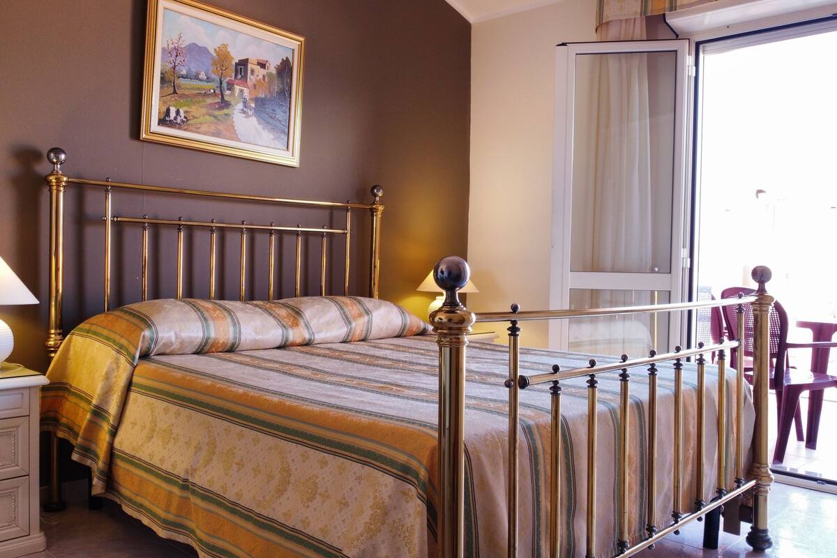 A neatly made bed with a striped bedspread, a bedside lamp, a nightstand with drawers, a painting on the wall, and a view of the outdoors through a window.