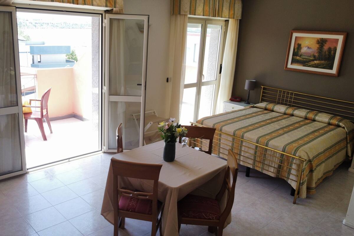 A hotel room with a bed, a table with chairs, a small table with a vase of flowers, and a painting on the wall.