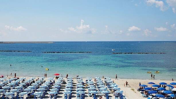 A beach with rows of blue umbrellas and chairs, a clear blue sea, and a few people in the water. - Hotel San Marco (Alghero)