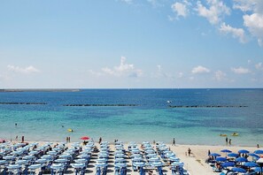 Una playa con hileras de sombrillas y sillas azules, un mar azul cristalino y algunas personas en el agua.