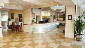 A reception area with a checkered floor, a reception desk, a seating area with chairs, and a potted plant. - Hotel San Marco (Alghero)