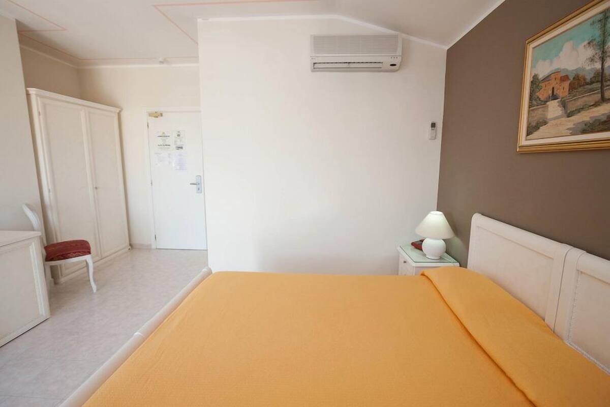 A bedroom with a bed, a nightstand with a lamp, an air conditioning unit, a door, and a painting on the wall.