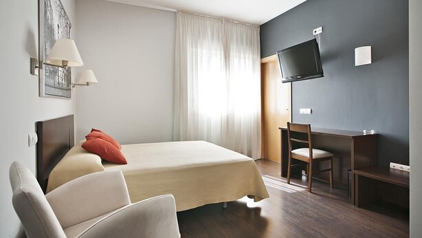 Habitación de hotel con cama, escritorio, silla, televisor y ventana con cortinas.