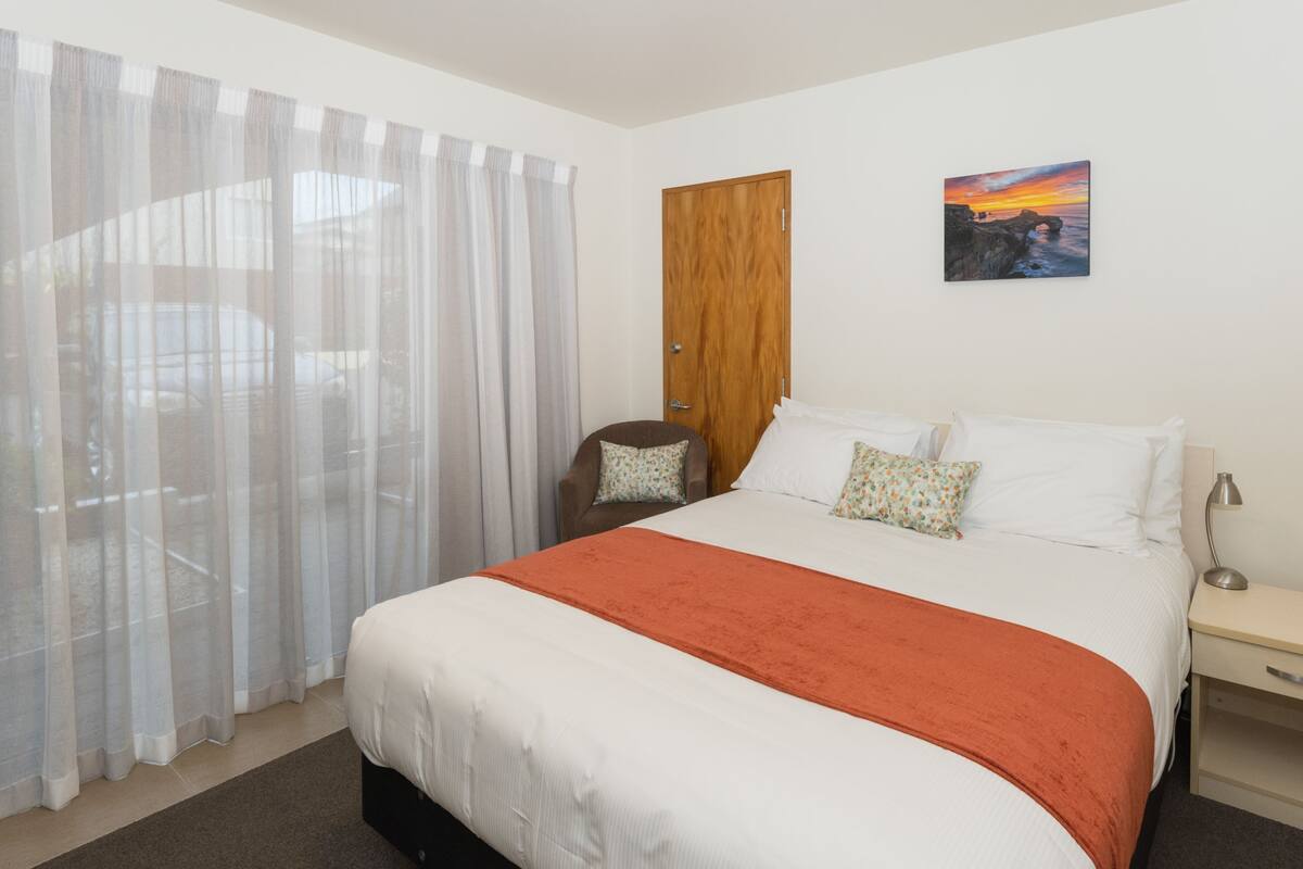 A bedroom with a bed, a nightstand, a chair, a wooden door, and a painting on the wall.