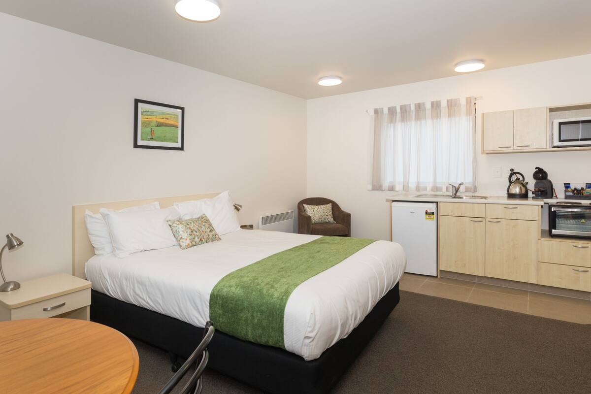 A hotel room with a bed, a desk, a chair, a kitchenette, and a window with curtains.