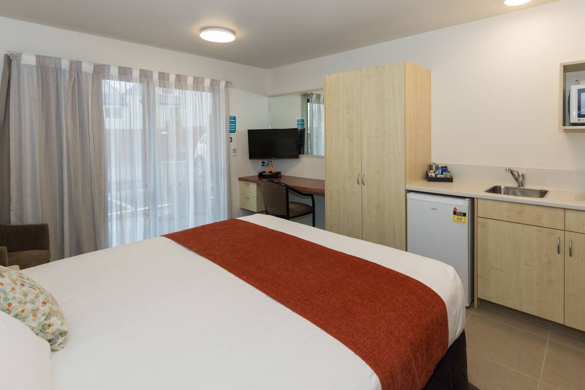 A hotel room with a bed, a desk, a television, a small kitchenette, and a bathroom.