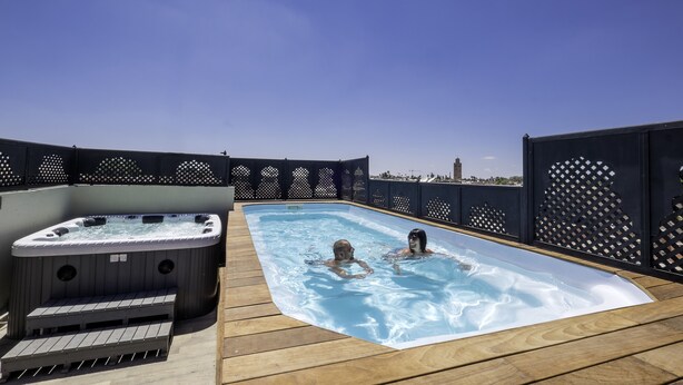 A hot tub and a swimming pool on a wooden deck with a view of a distant tower. - Riad Nesma Suites & SPA (Marrakech)