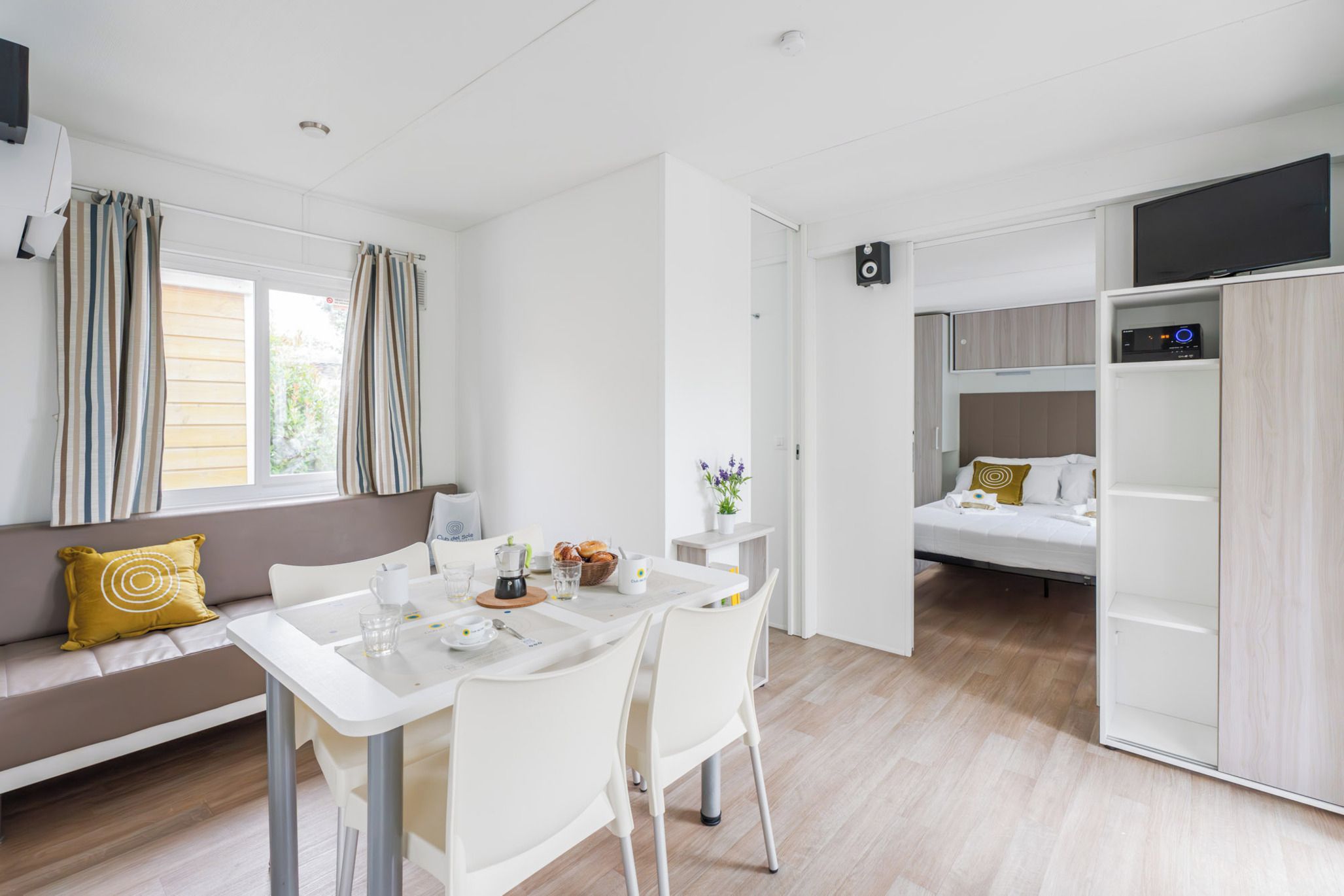 Une pièce moderne et bien éclairée, comprenant une table à manger, des chaises, un canapé avec des coussins, un lit et un téléviseur fixé au mur.
