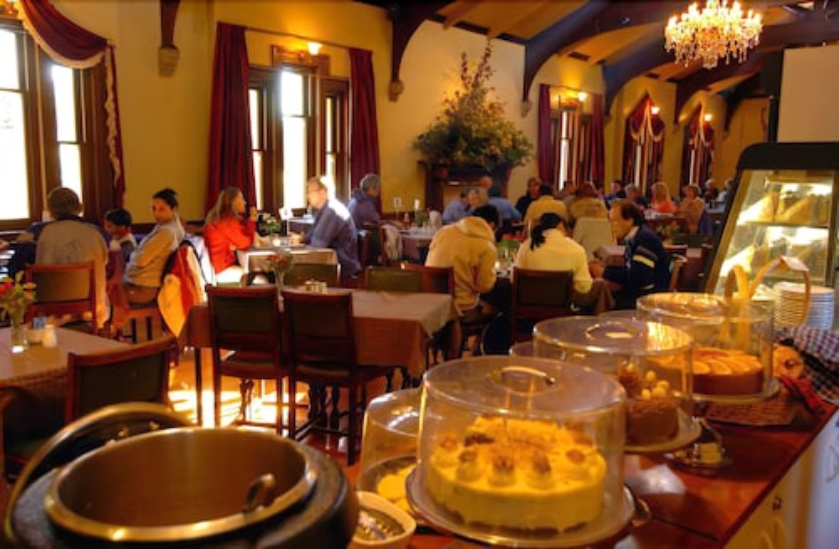 A restaurant with multiple tables and chairs, a variety of dishes displayed in the foreground, and a chandelier hanging from the ceiling.