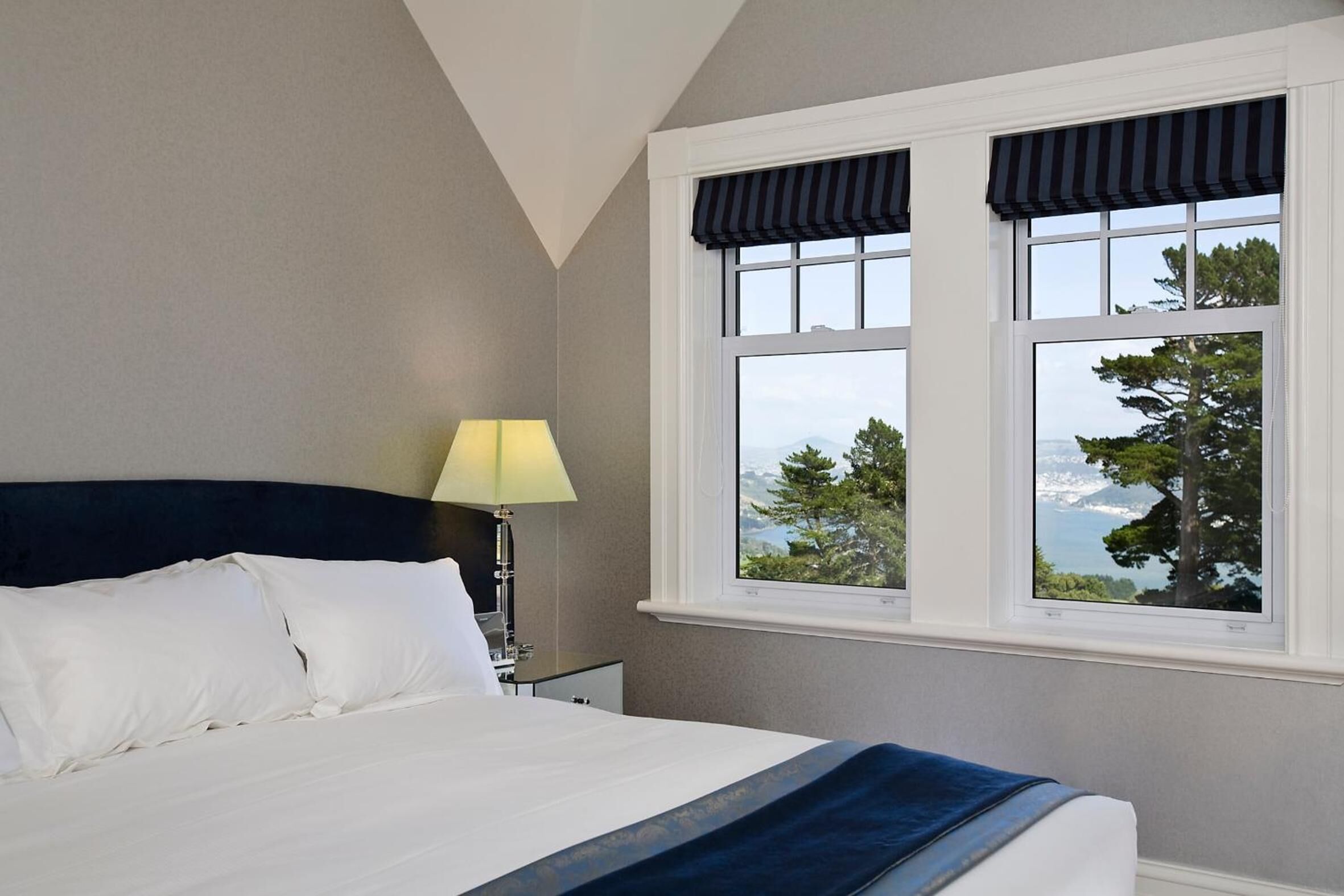 A neatly made bed with white linens, a bedside lamp, and a window with a view of trees and the sea.
