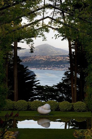 Un jardin paisible avec un bassin aux eaux calmes, entouré de grands arbres et offrant une vue sur une ville lointaine et des montagnes.