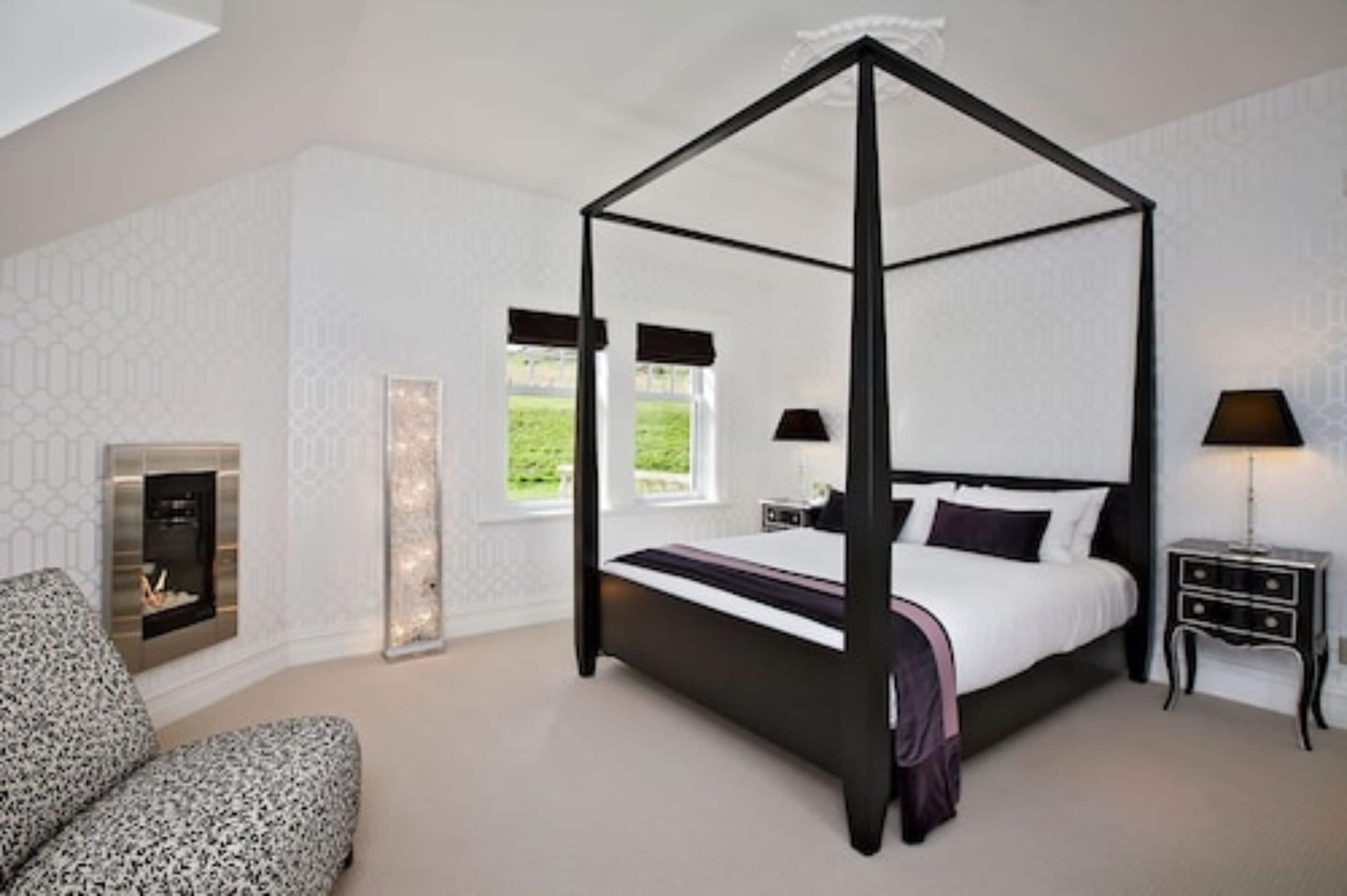 A four-poster bed in a bedroom with a fireplace, a nightstand, and a chair.