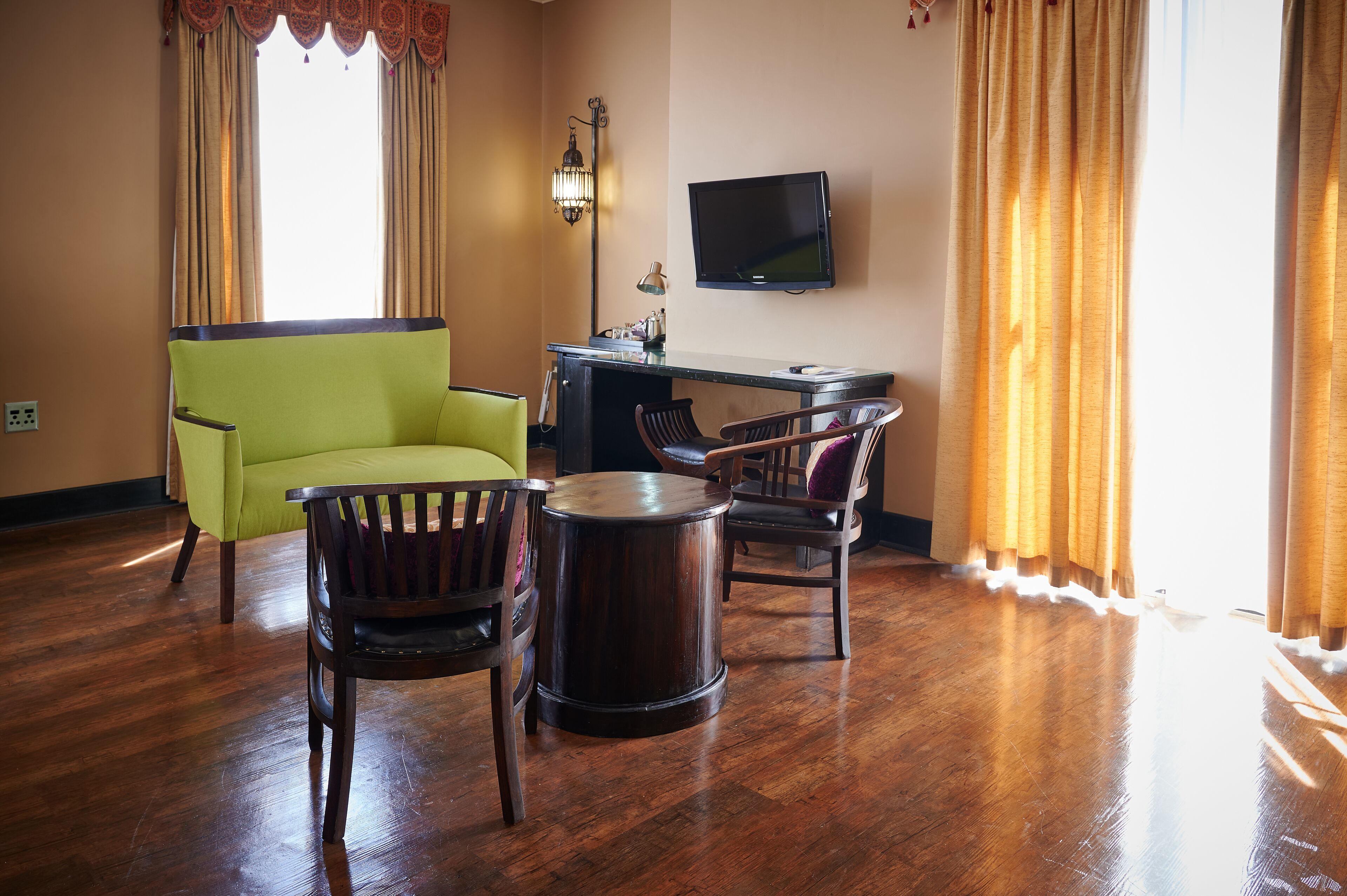 A room with a wooden floor, a green armchair, a round table, and a desk with a chair. A flat-screen TV is mounted on the wall.