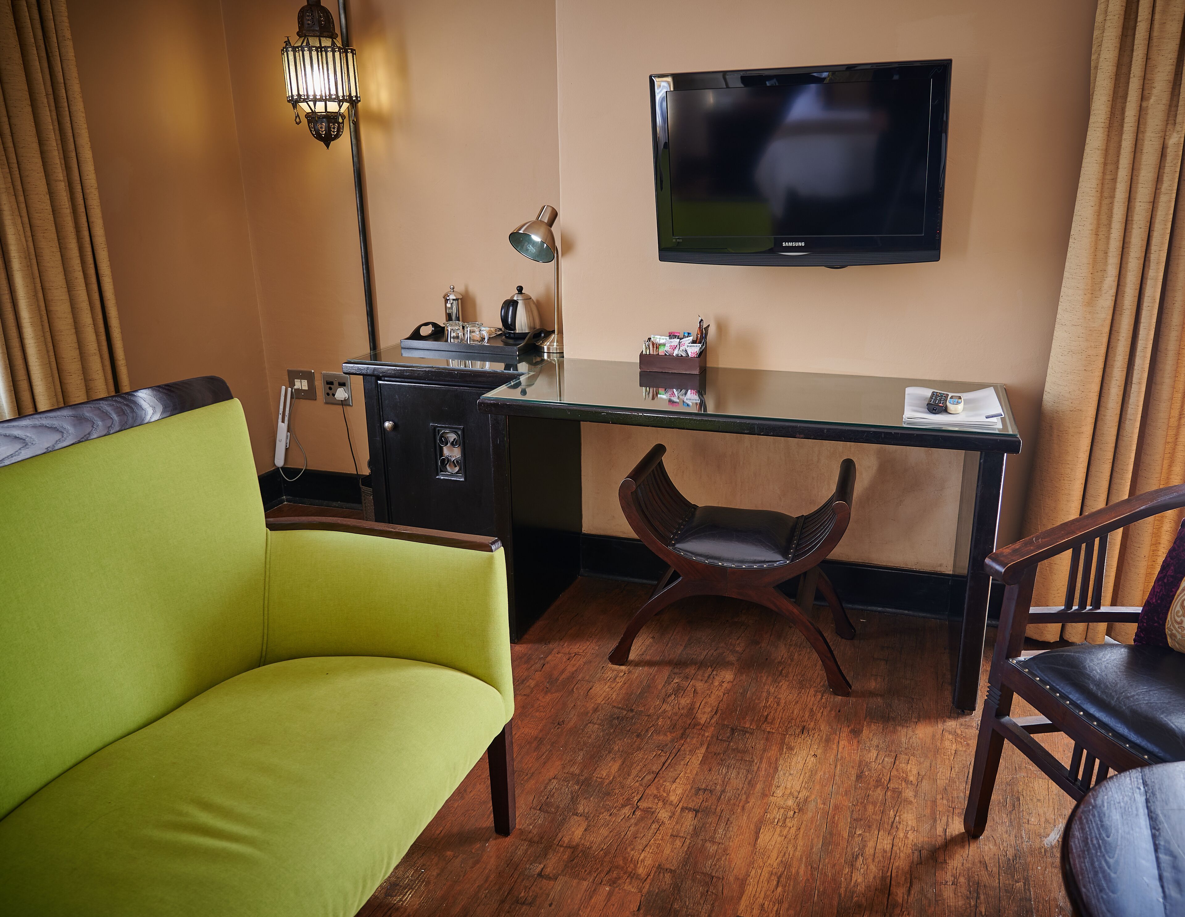 Une chambre d’hôtel avec un canapé vert, une chaise en bois, un bureau avec un ordinateur, une lampe et un téléviseur à écran plat fixé au mur.