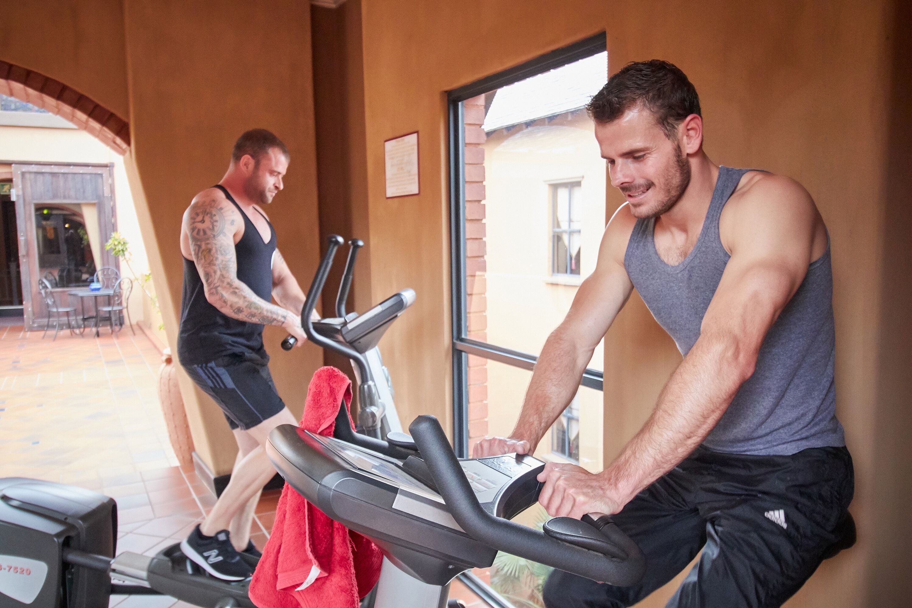 Two men exercising on stationary bikes in a gym with a brick wall and a window.
