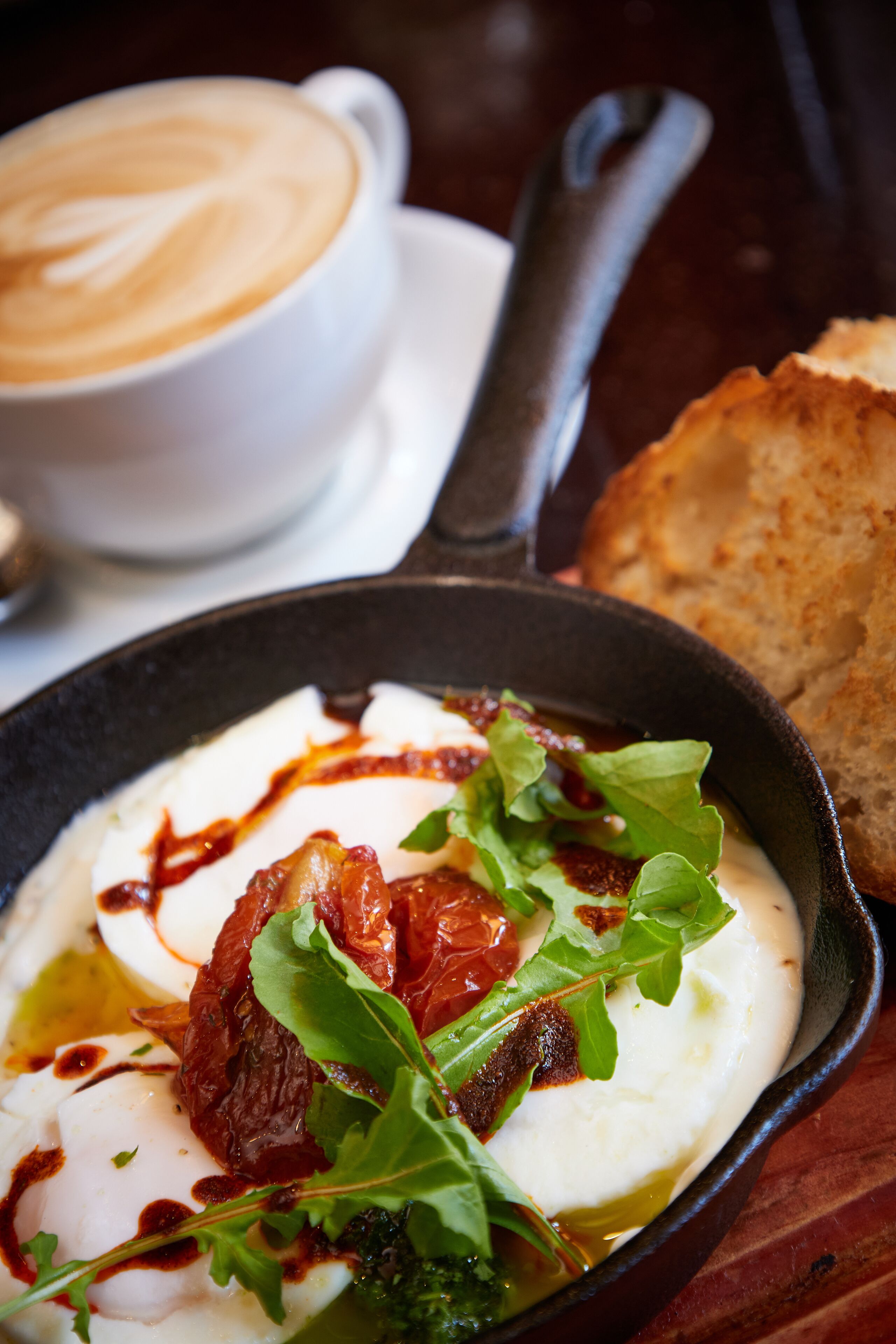 A breakfast setting with a white plate of eggs, greens, and sun-dried tomatoes, a cup of coffee, and a piece of toast on a wooden table.