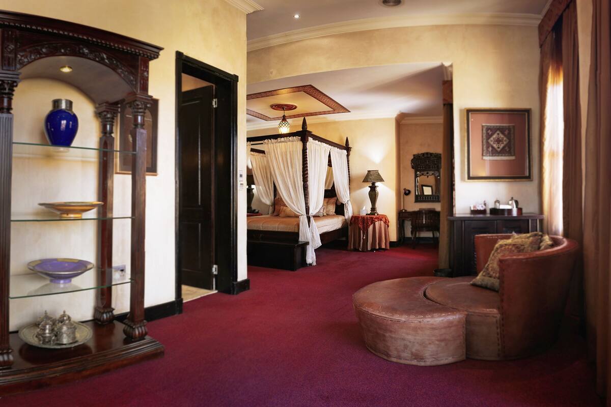 A bedroom with a four-poster bed, a sitting area with a chair and ottoman, a glass display cabinet, and a red carpet.