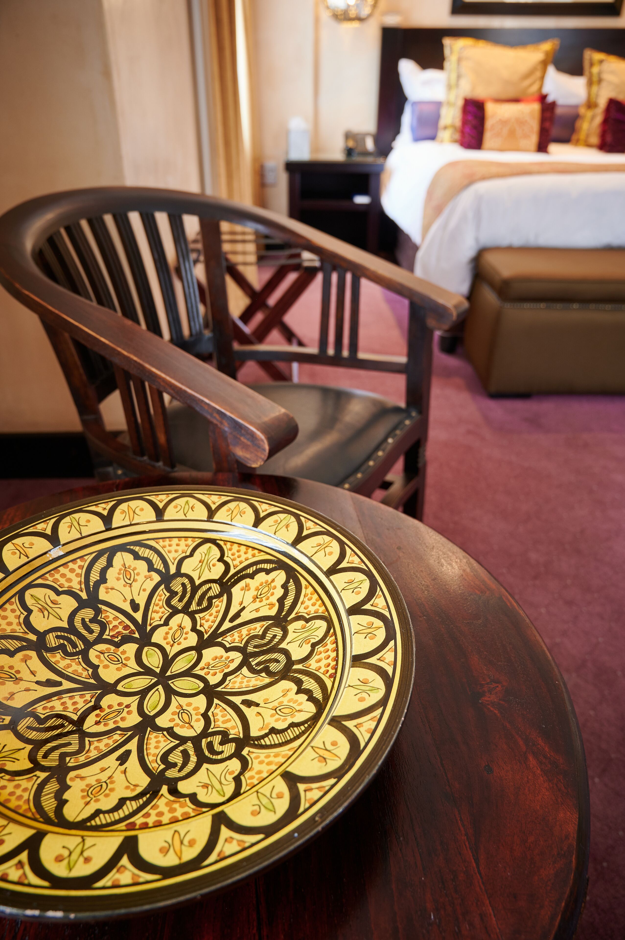 A wooden chair with a patterned cushion, a round wooden table, and a bed with pillows in the background.