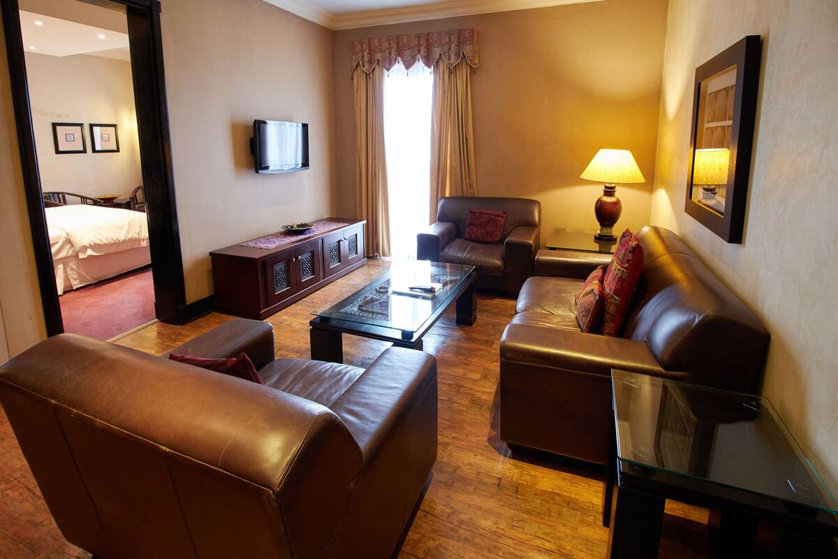 A hotel room with a living area featuring leather sofas, a glass coffee table, a television mounted on the wall, and a bed in the background.