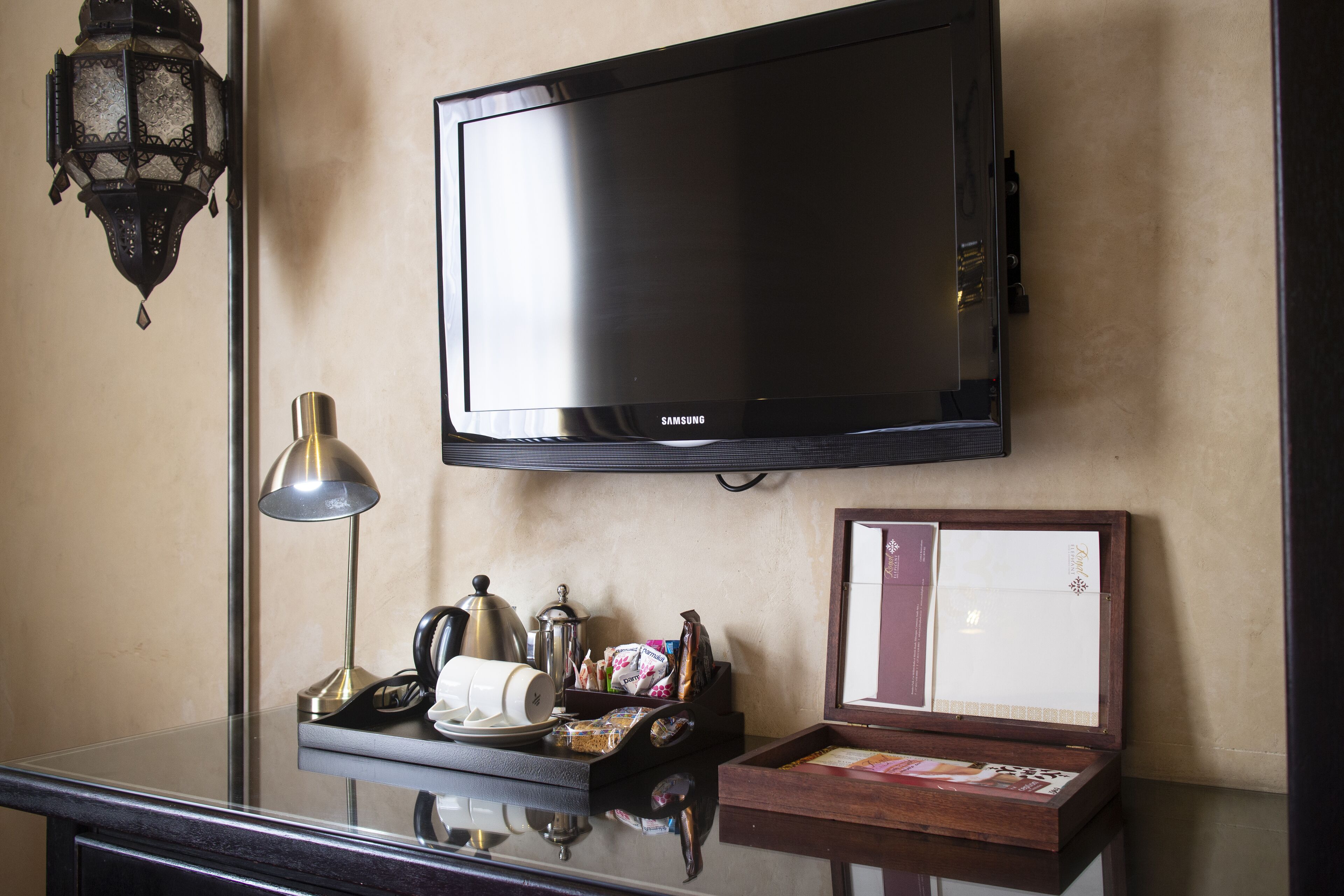 Une chambre d’hôtel équipée d’un téléviseur à écran plat fixé au mur, d’une table de chevet avec une lampe, d’une machine à café et d’un plateau avec des en-cas et une carte.