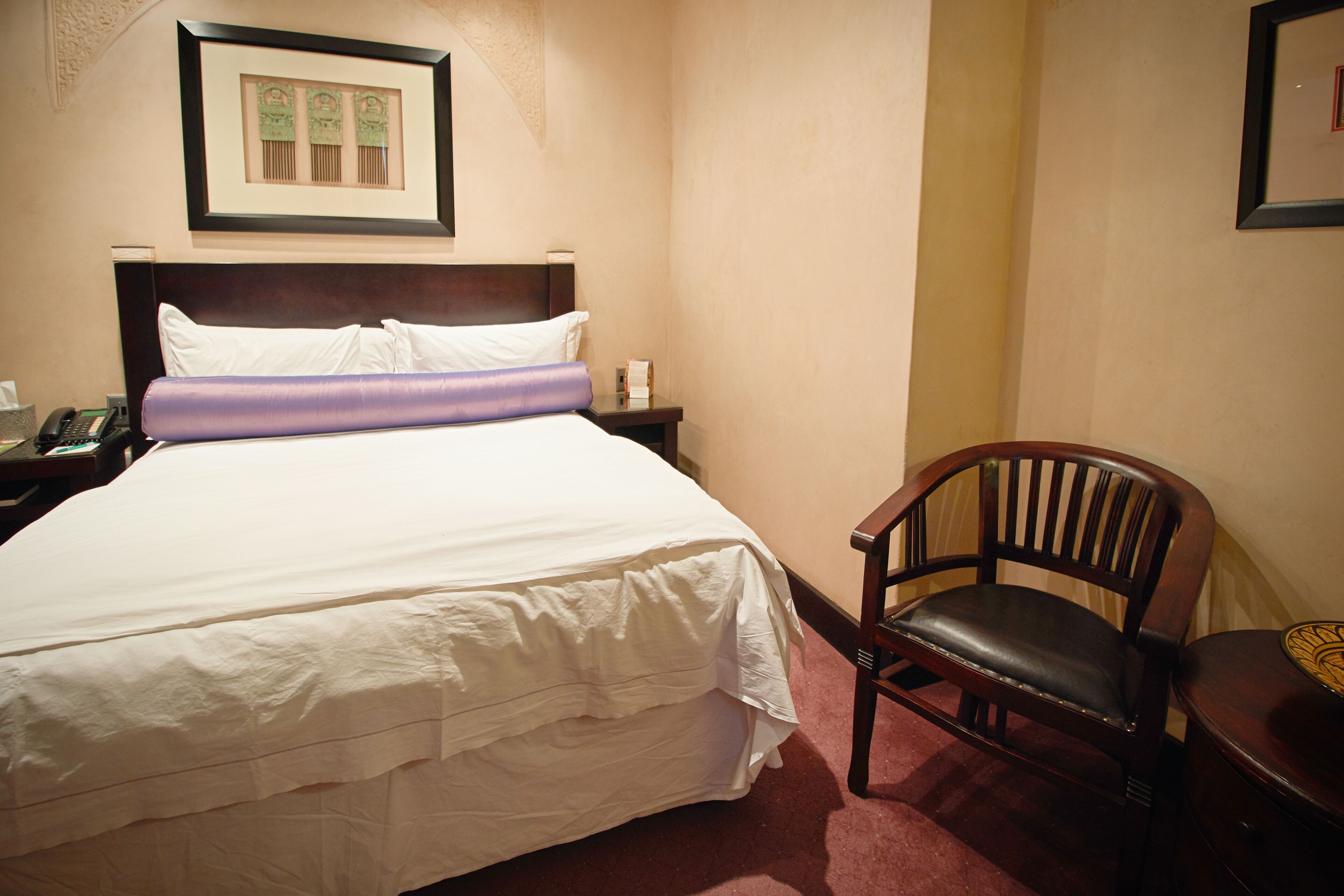 Een hotelkamer met een groot bed, een houten stoel, een nachtkastje en een ingelijst kunstwerk aan de muur.