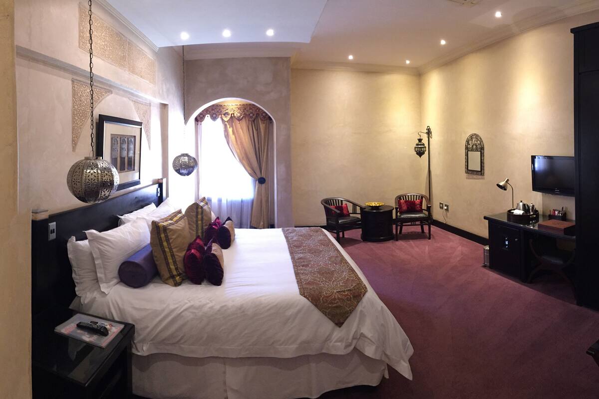 A spacious hotel room with a large bed, two armchairs, a desk with a TV, and a decorative lamp.