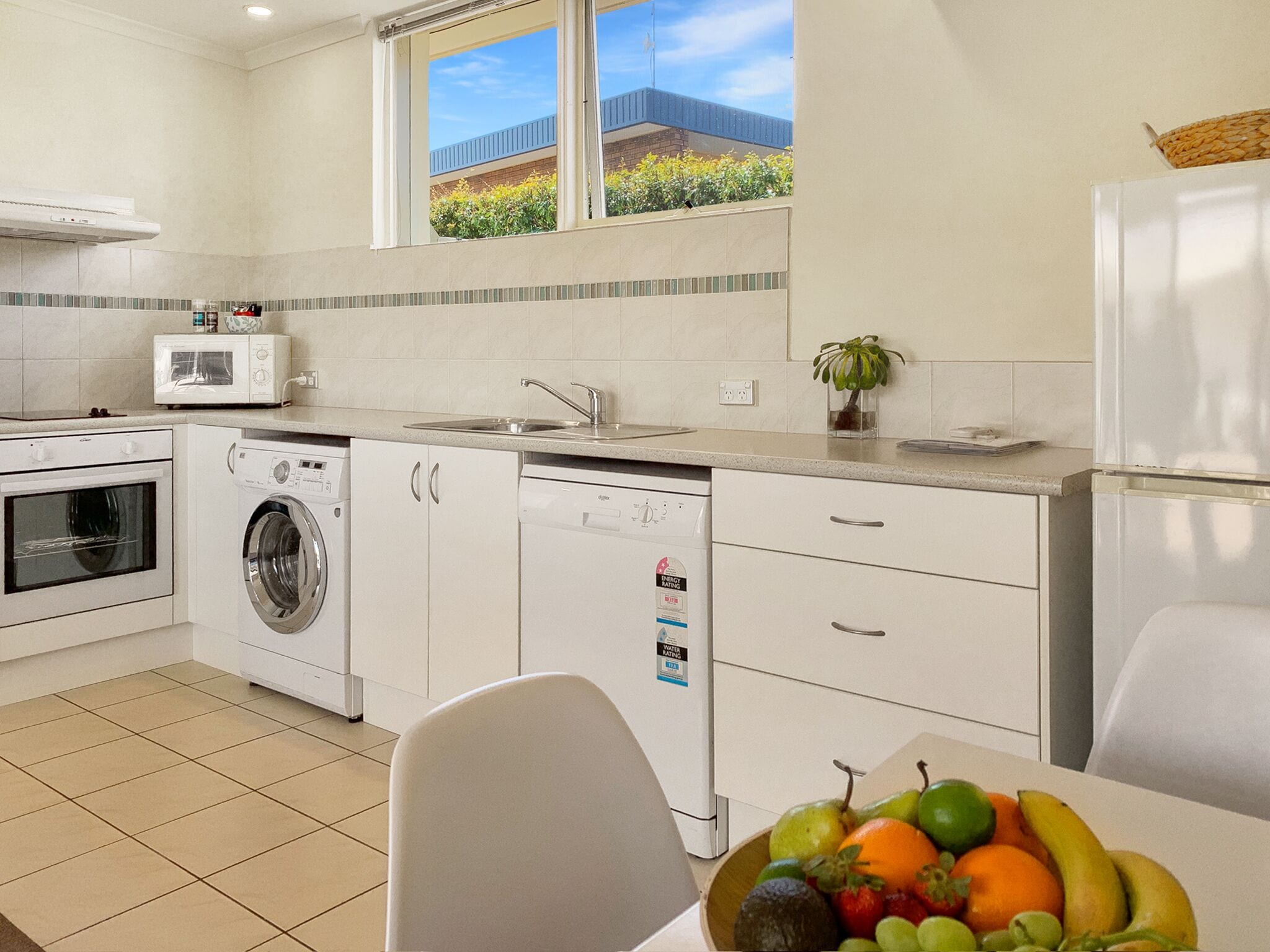 Une cuisine moderne équipée d’un lave-linge, d’un lave-vaisselle et d’un four à micro-ondes. Une fenêtre offre une vue sur les bâtiments et la végétation environnante.