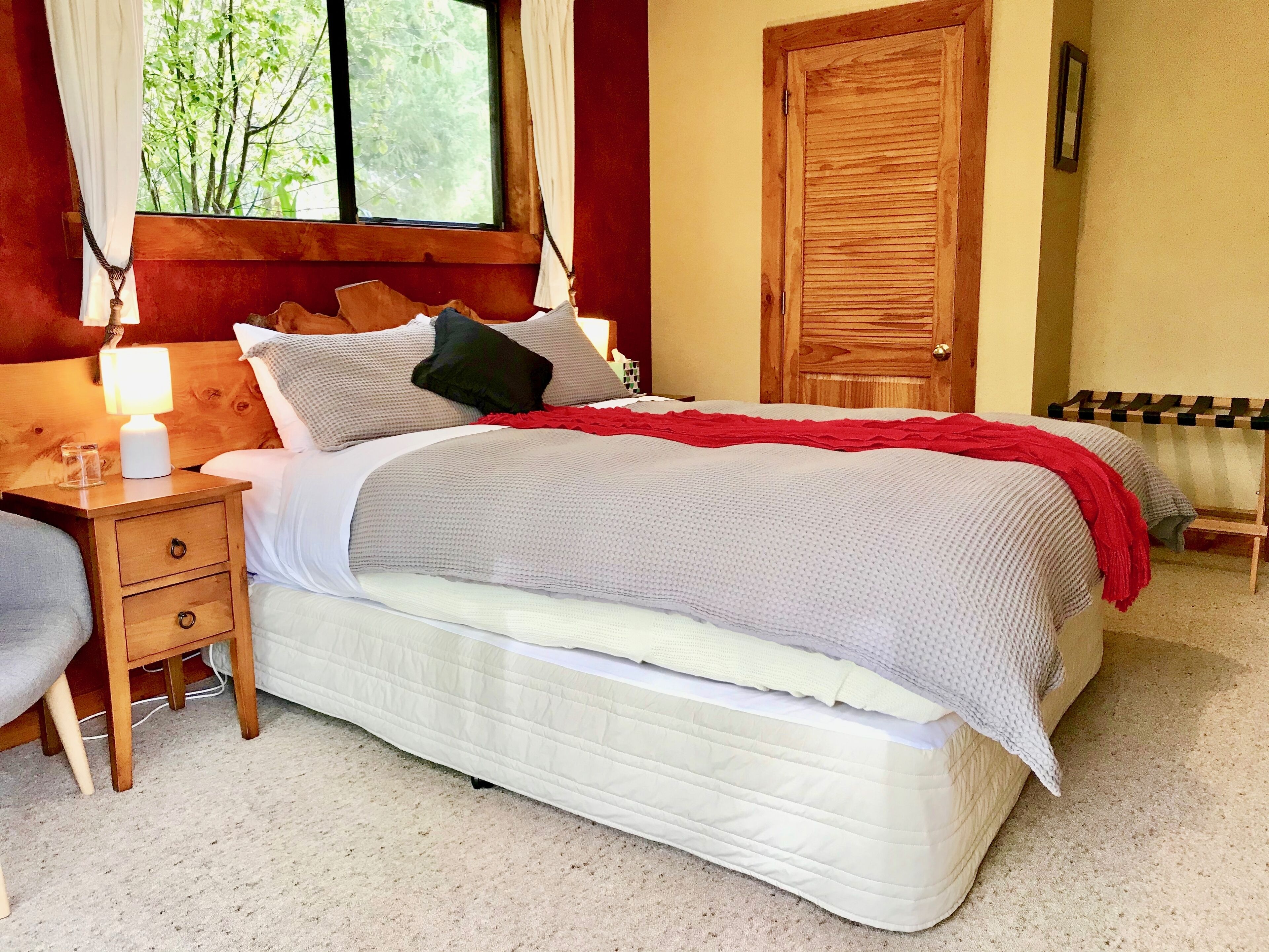 A neatly made bed with a white mattress, gray and red bedding, and a wooden headboard. There is a wooden nightstand with a lamp and a mirror on the wall.