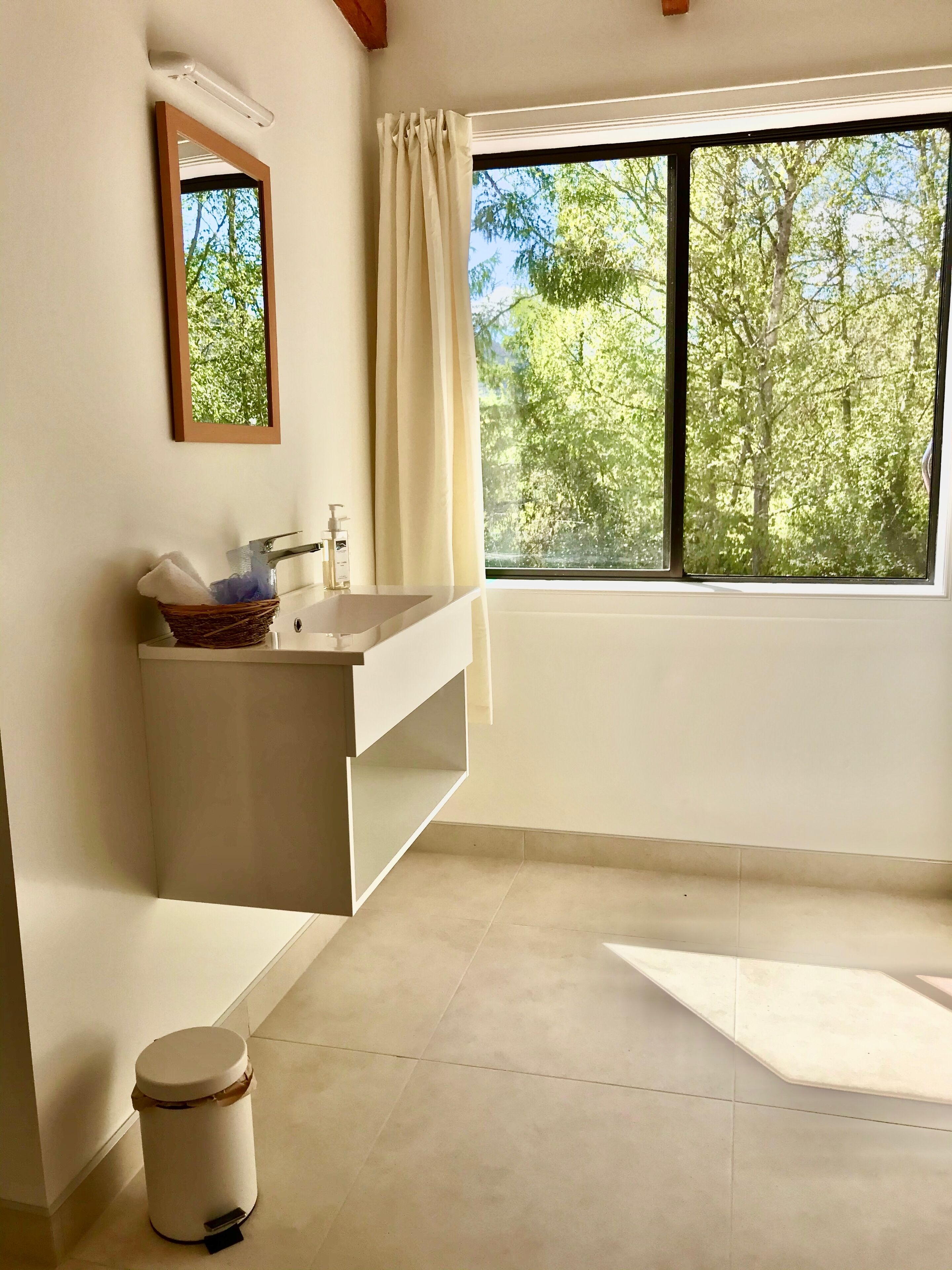 Une salle de bain avec une grande fenêtre, un lavabo avec un miroir et une poubelle.