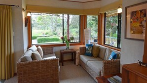 A cozy room with wicker furniture, a small table with a vase, and large windows offering a view of greenery. - Murchison Lodge (Murchison)