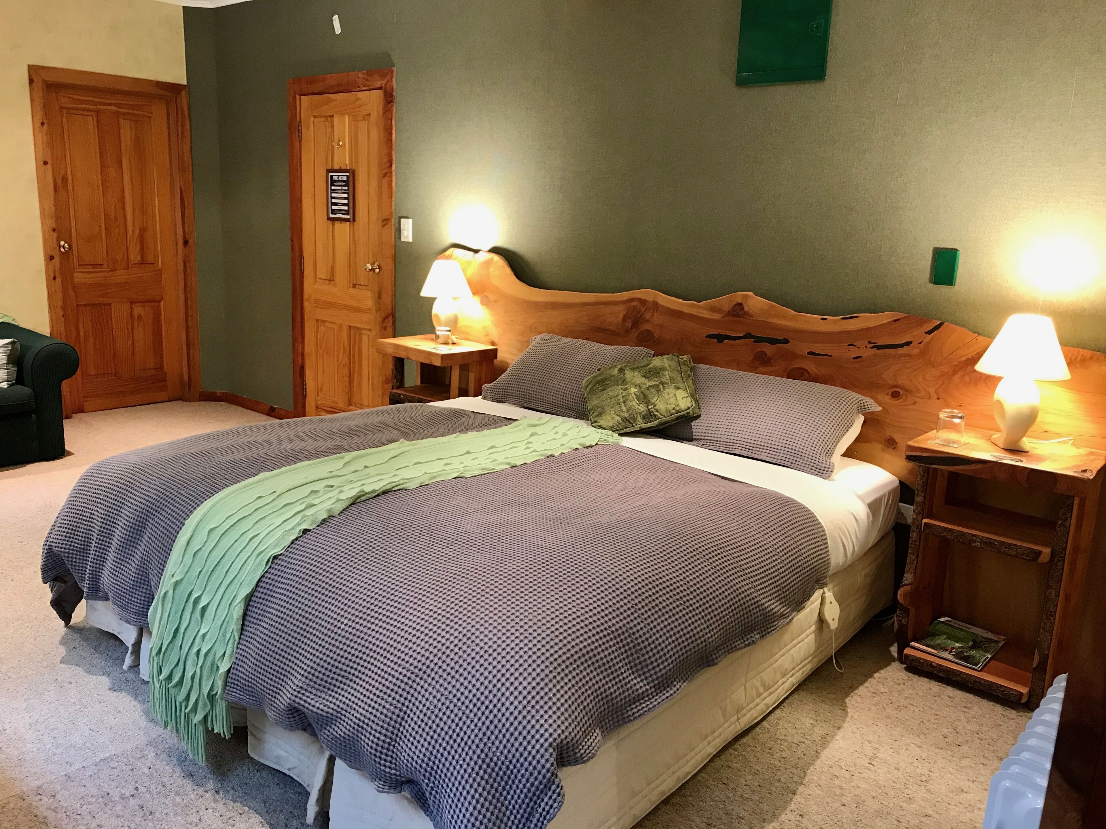 Una cama bien tendida con un plaid verde, dos lámparas de noche y un cabecero de madera.