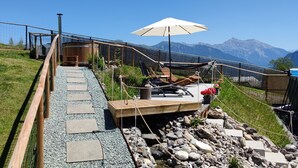 A wooden deck with stone steps leading to a seating area with lounge chairs and a white umbrella. - Gasthaus Alpina (Tschappina)
