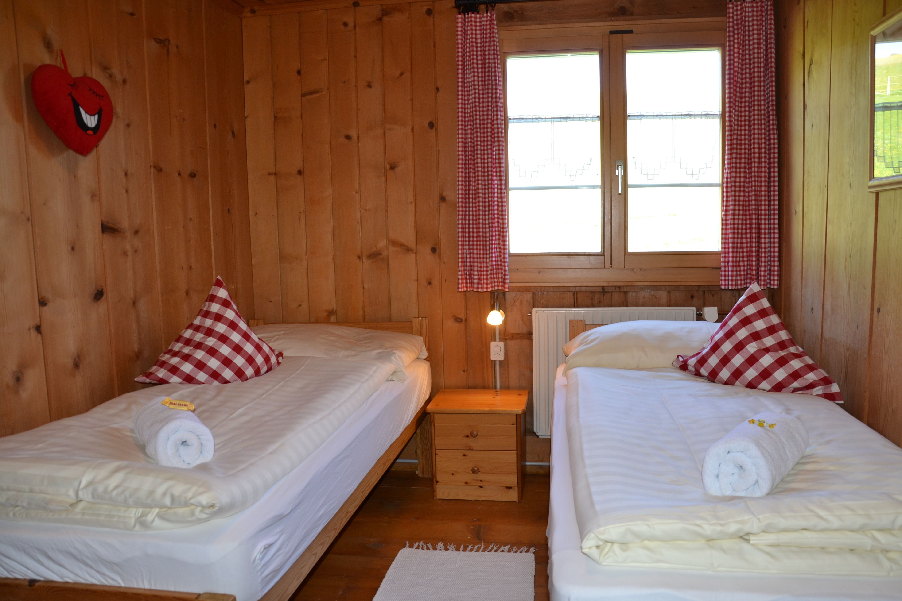 A wooden cabin room with two beds, each with a red and white checkered pillow, a heart-shaped decoration, and a lamp between the beds.