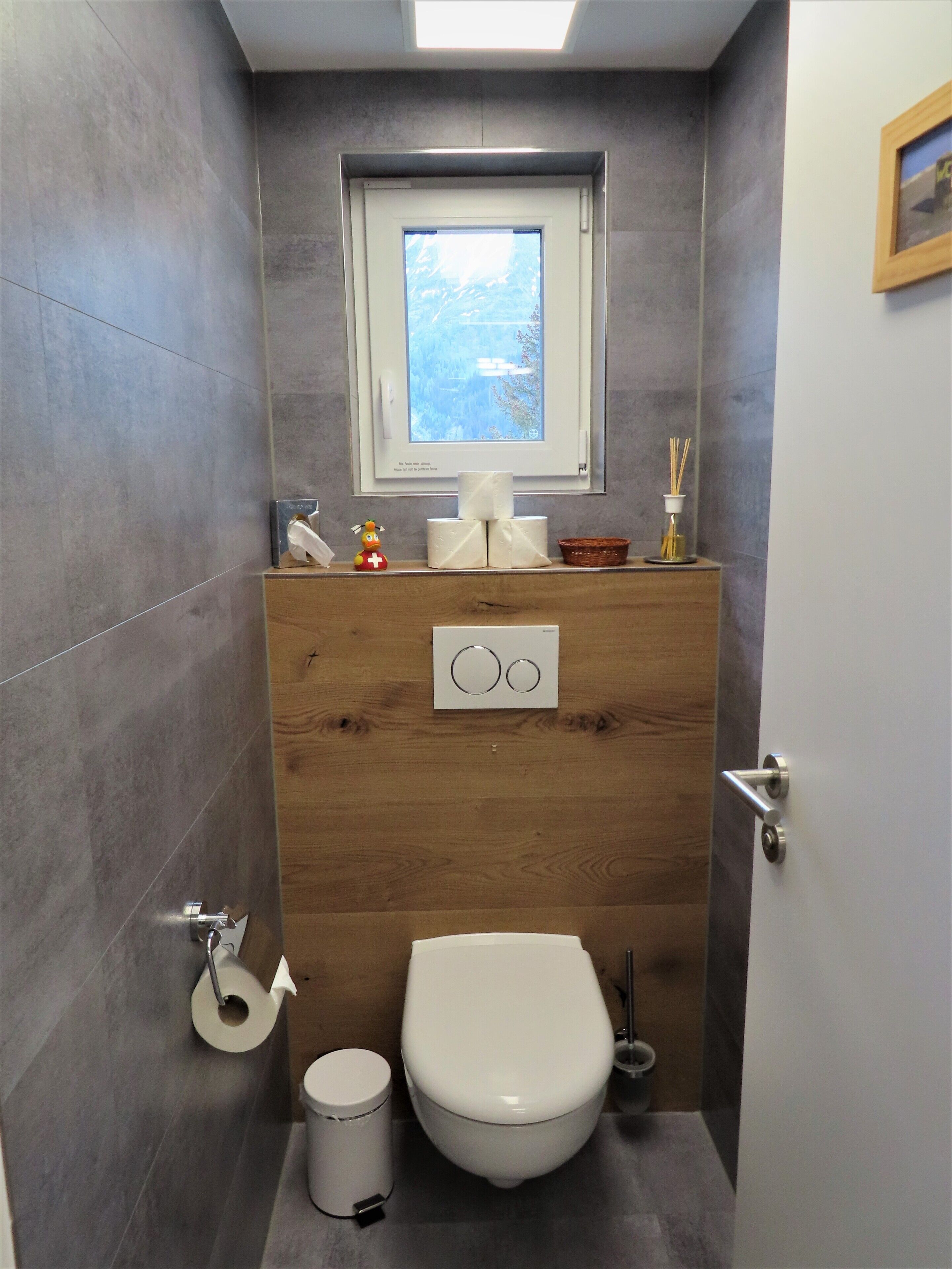 Een compacte badkamer met toilet, een klein raam en een houten wastafelmeubel met wastafel.