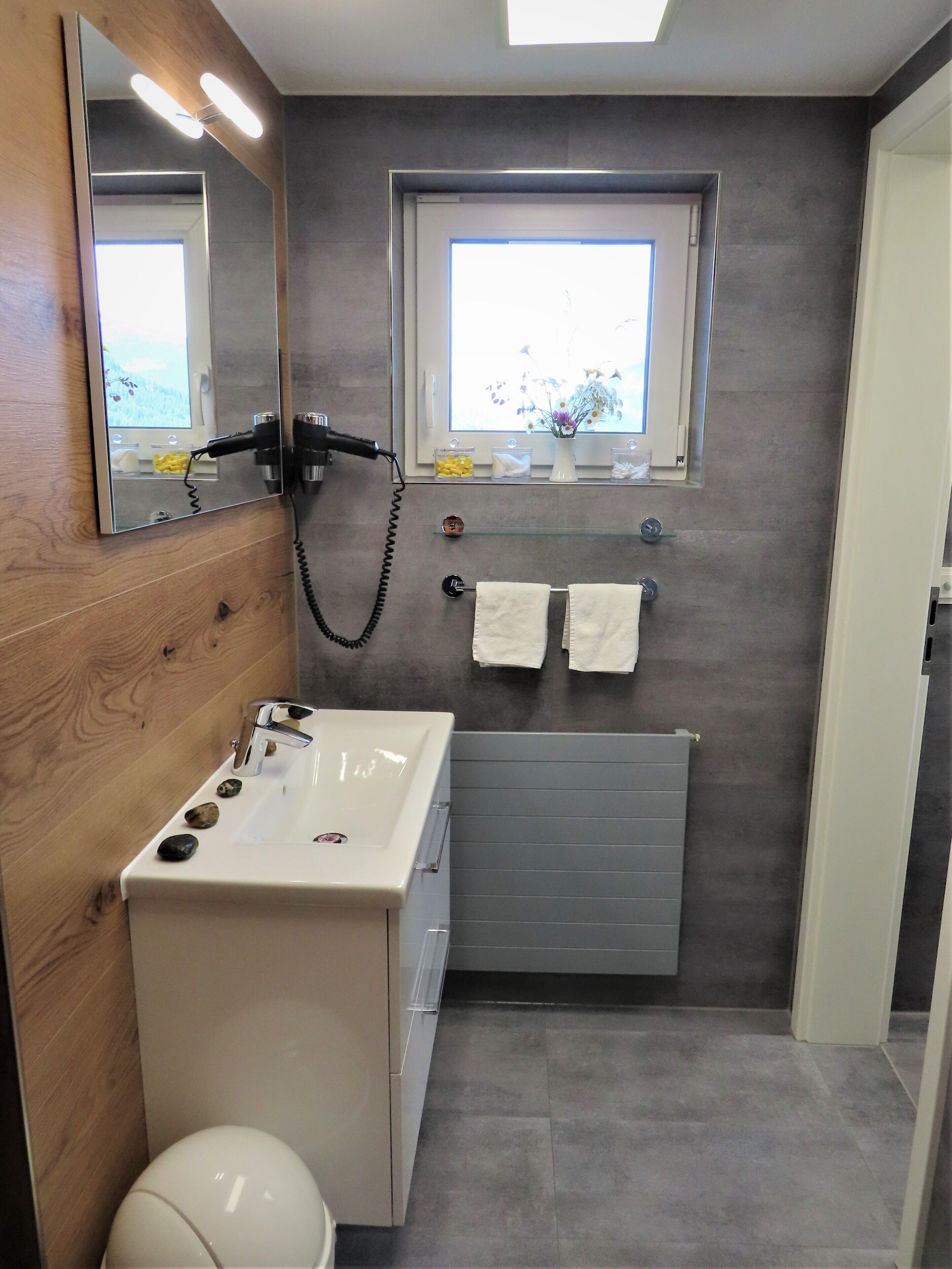 Une salle de bain avec un lavabo blanc, un miroir, un porte-serviettes, une fenêtre donnant sur un paysage agréable et des toilettes.