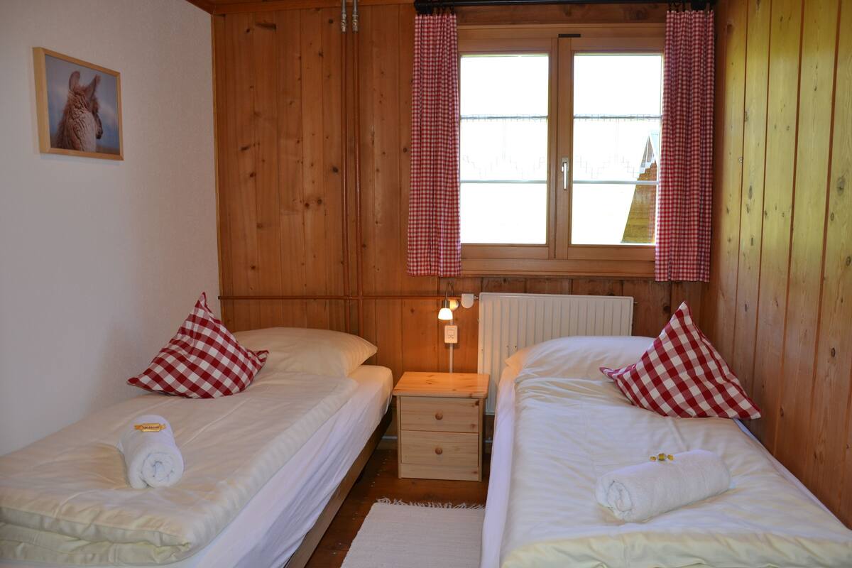 A wooden cabin room with two beds, checkered curtains, a small table, and a radiator.