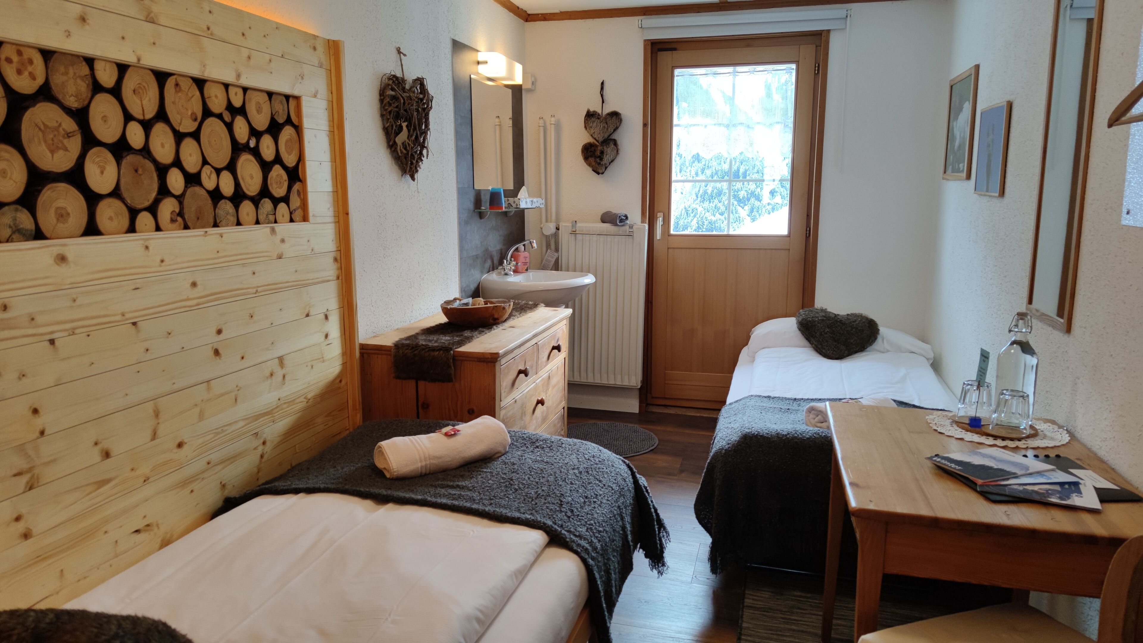 A cozy room with two beds, a wooden wall, a window, a sink, and a table with a bottle and glasses.