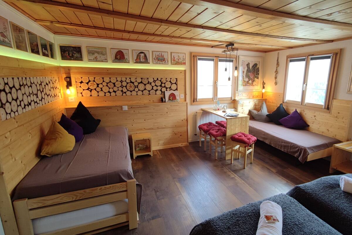 A cozy cabin-style room with a wooden ceiling, a bed with a brown mattress, a bench with cushions, and a small table with a lamp.