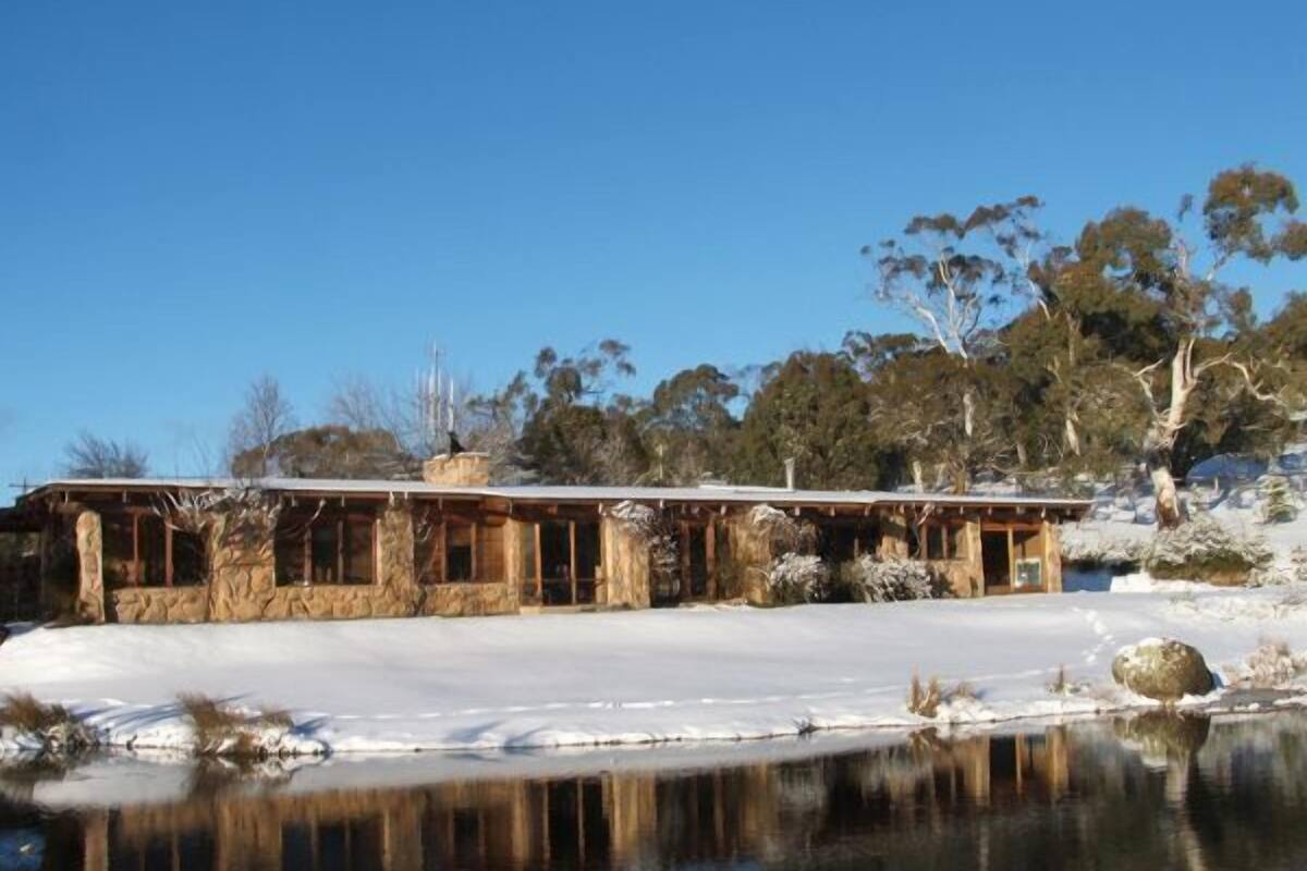 Un bâtiment rustique en pierre, doté de grandes fenêtres, situé au bord d’un plan d’eau aux reflets changeants, entouré d’arbres et sous un ciel dégagé.