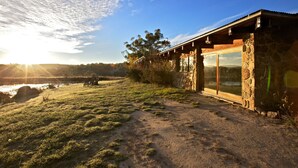 A stone building with large glass doors and windows, a grassy area with a bench, and a view of the sunset over a body of water. - Snowy Wilderness (Ingebirah)