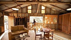A rustic cabin interior with a wooden floor, a sofa, a dining table, and a person sitting by the window. - Snowy Wilderness (Ingebirah)